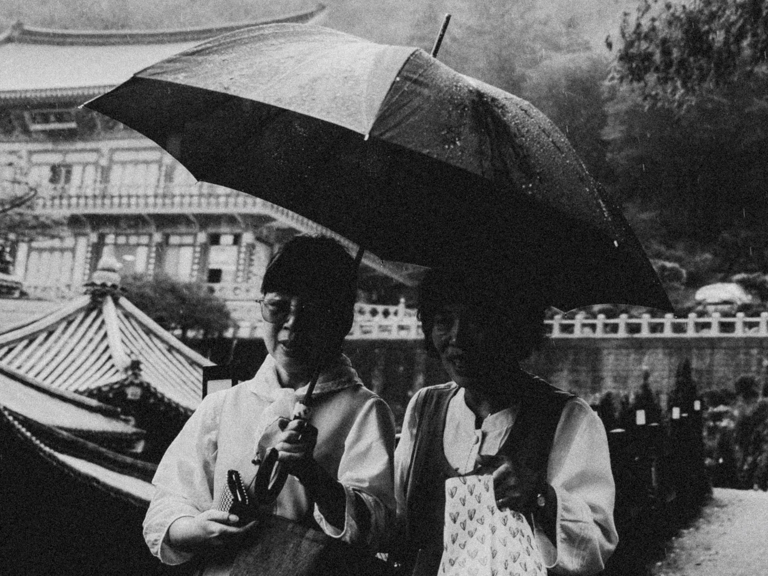 A black-and-white photo of a couple holding umbrellas and standing close together outdoors. The woman is holding a paper with heart shapes, and both are dressed casually. In the background, there is a large amusement park ride and a building.