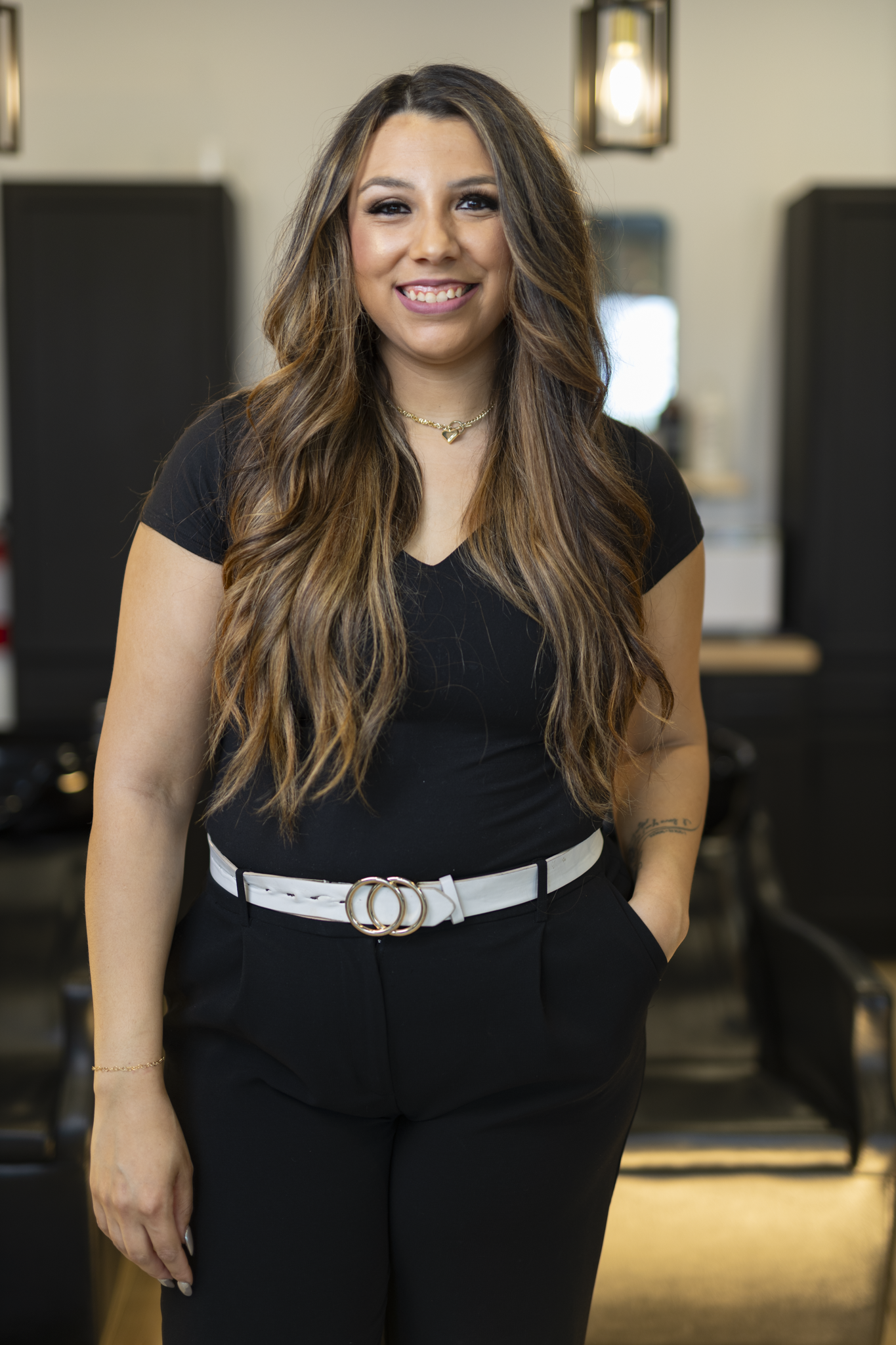 A woman with long, wavy brown hair and a bright smile is standing indoors, wearing a black top, black pants with a white belt, and accessorized with a gold necklace and bracelet.