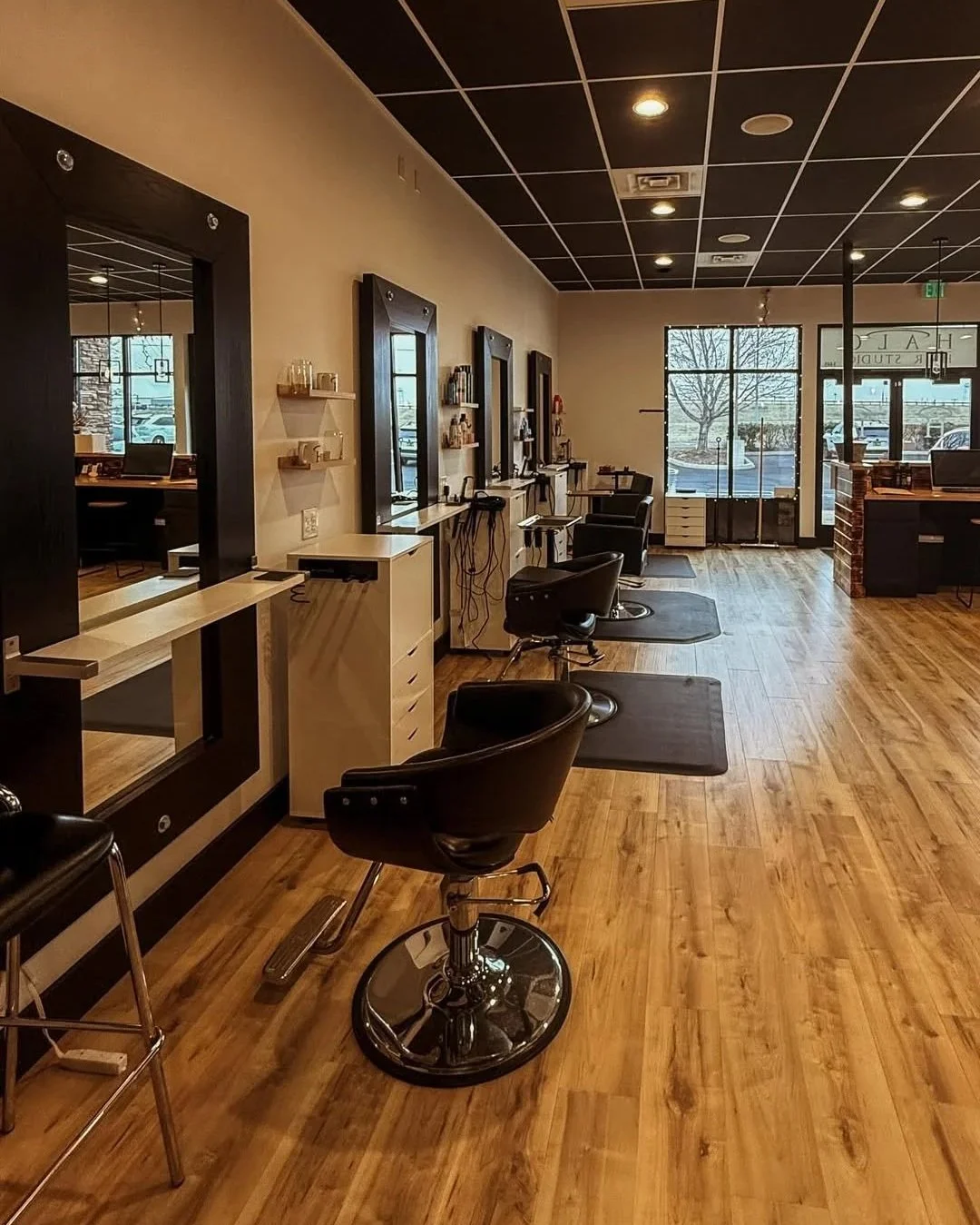 Empty hair salon with black chairs, mirrors, wooden flooring, and large front window showing parking lot outside.