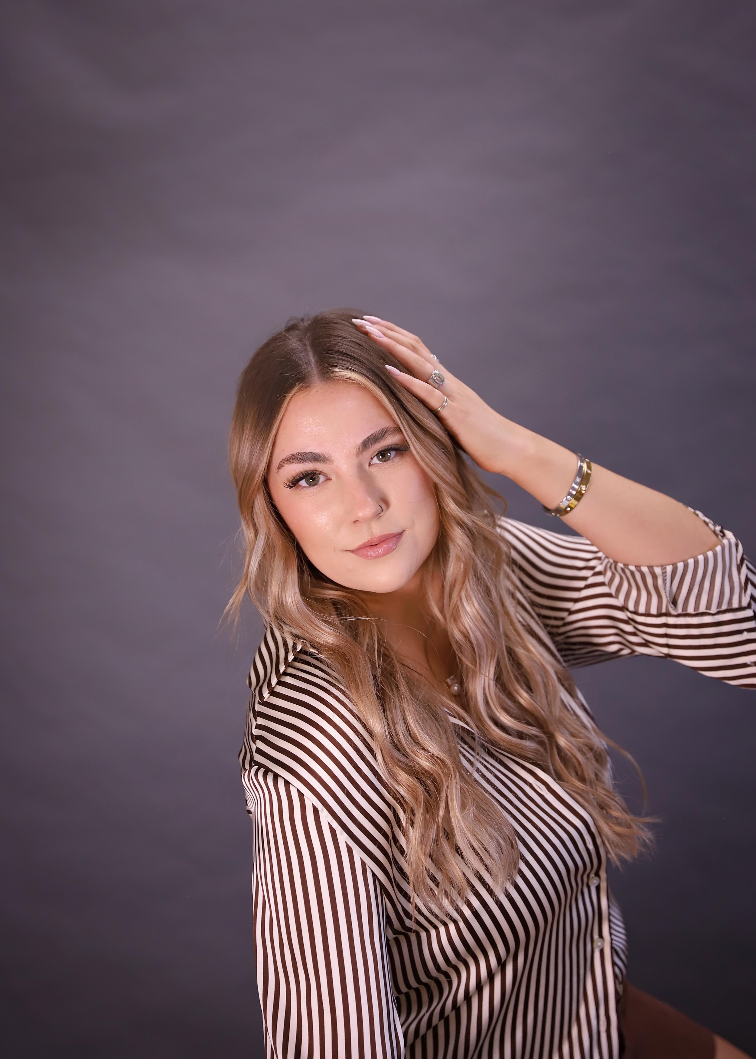 A young woman with long, wavy blonde hair, wearing a striped blouse, posing with her right hand on her head against a gray background.