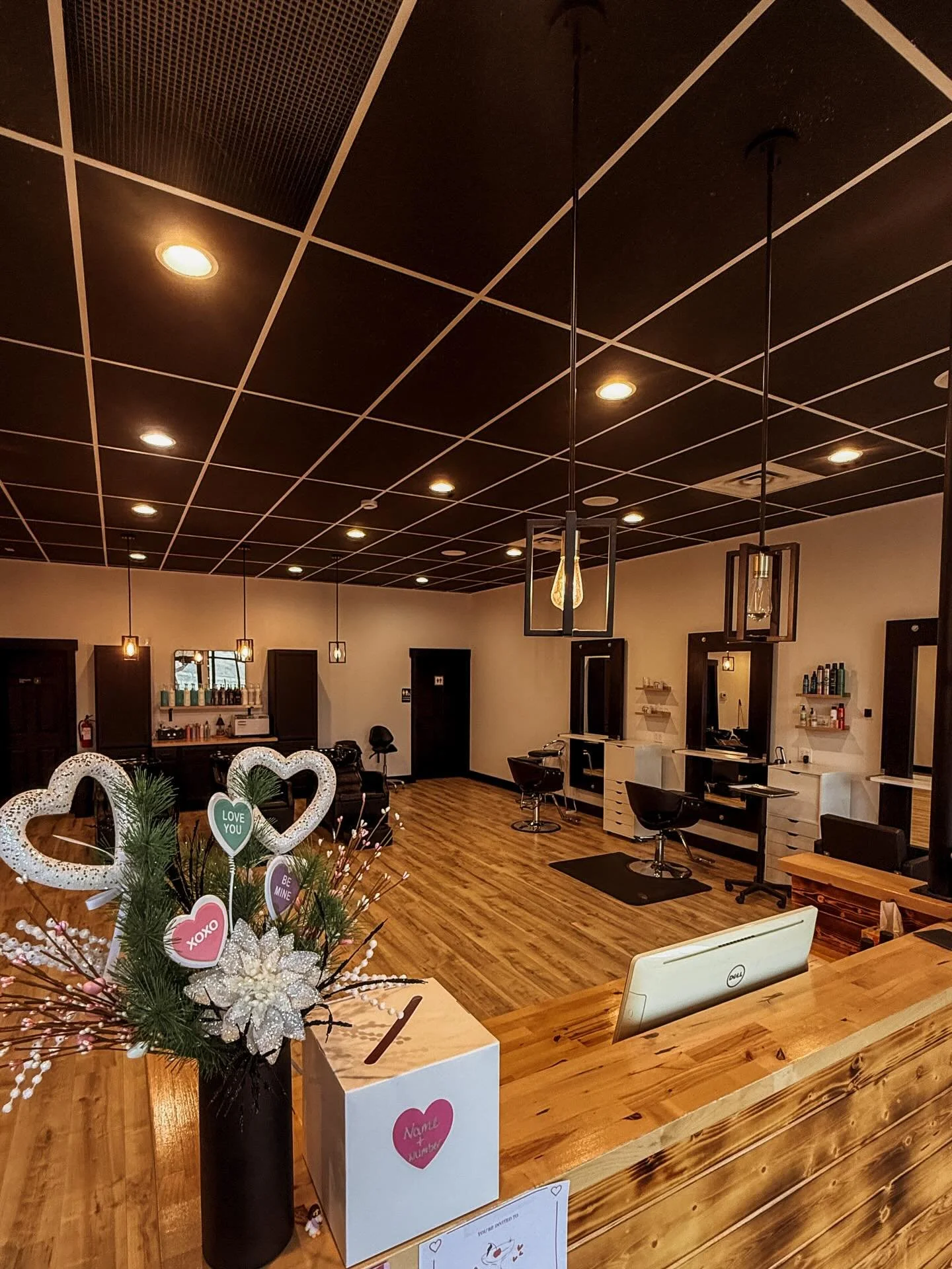 Empty hair salon with wooden floors, black ceiling with lighting, mirrors, chairs, and a reception desk decorated with heart-shaped and love-themed ornaments.