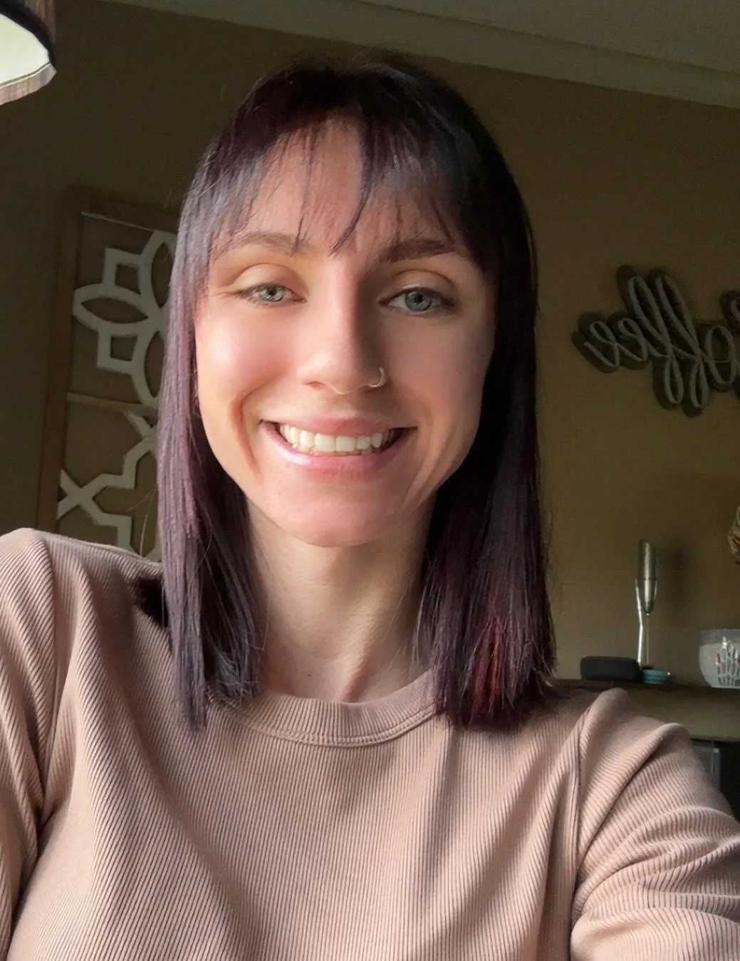 A woman with shoulder-length dark hair, light skin, and a nose ring smiling for a selfie in a home setting.