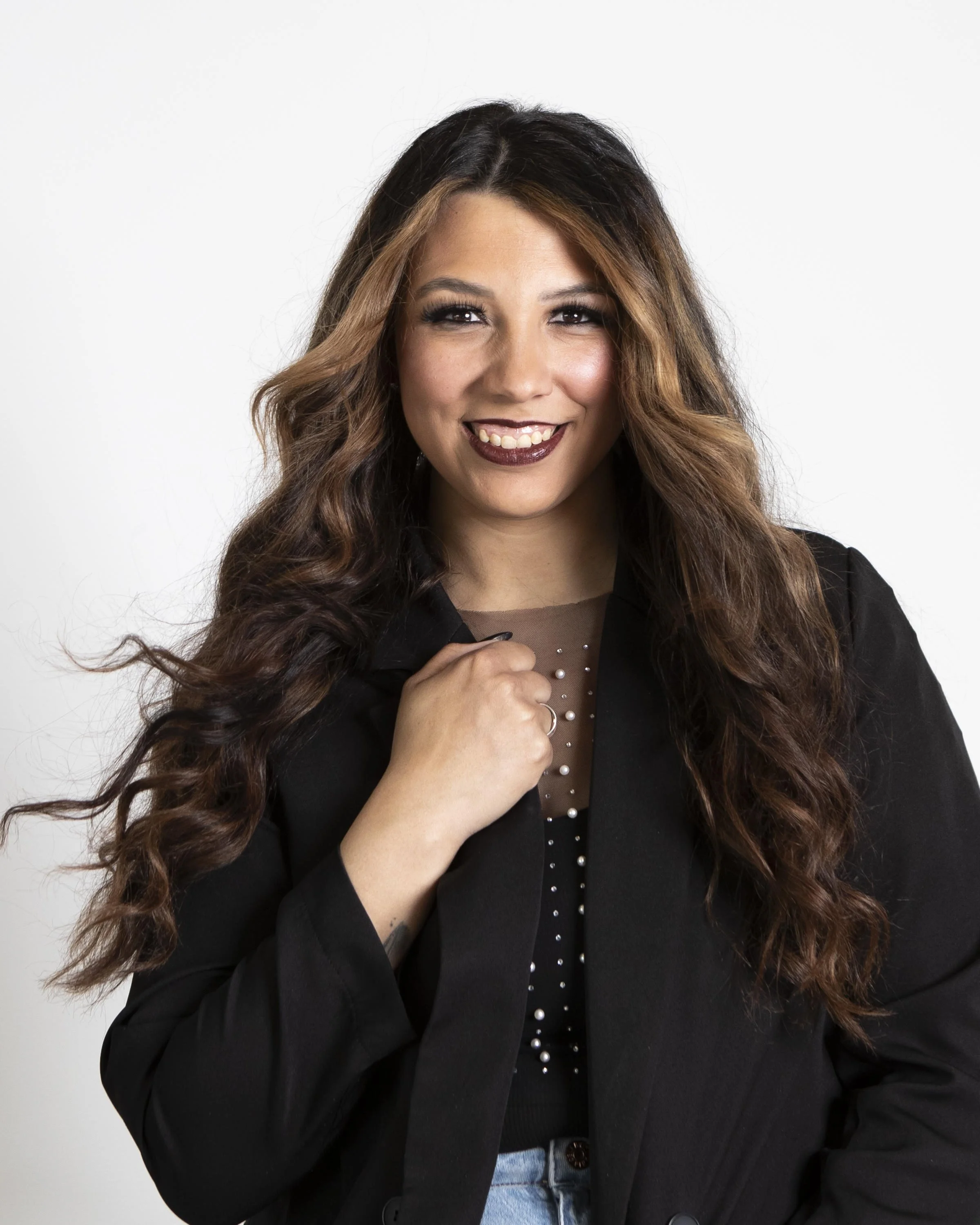 A woman with long, wavy brown hair and dark makeup, smiling and wearing a black jacket over a sheer top with pearls, standing against a white background.