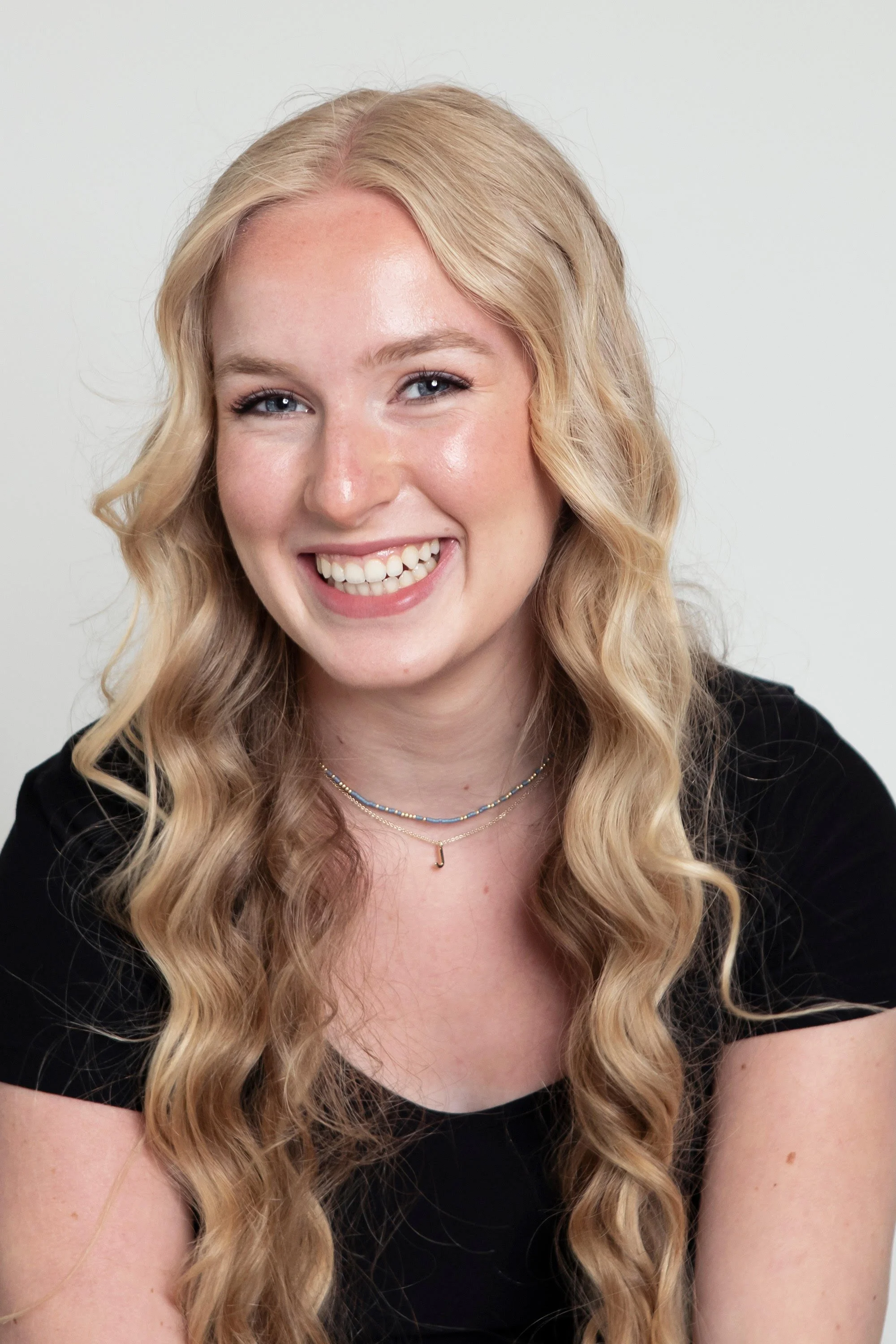 Young woman with long blonde wavy hair smiling at camera, wearing a black top and layered necklaces, against a plain light background.