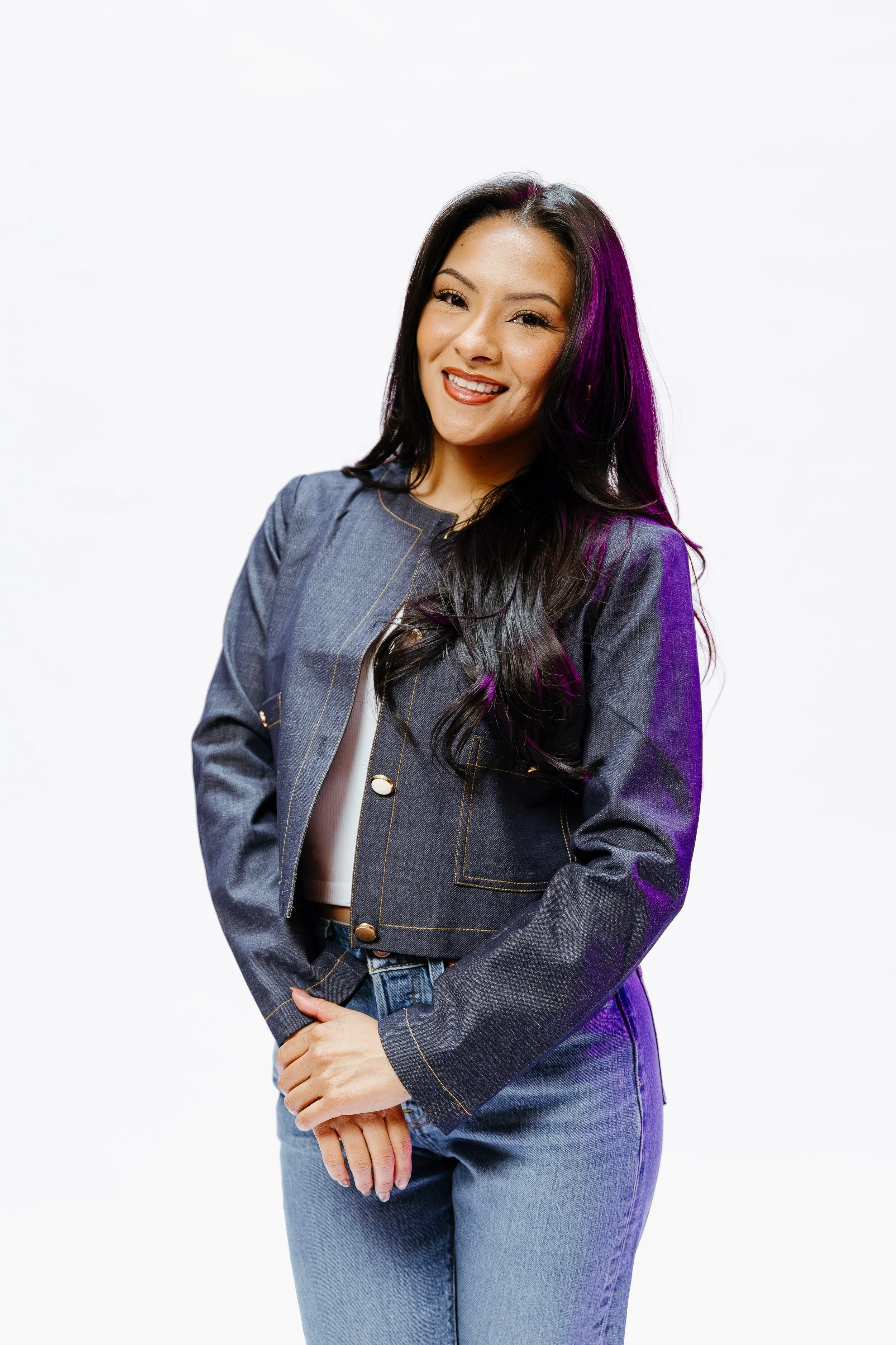 A young woman with long black hair, some purple highlights, smiling and looking at the camera, wearing a denim jacket and jeans, standing against a plain white background.