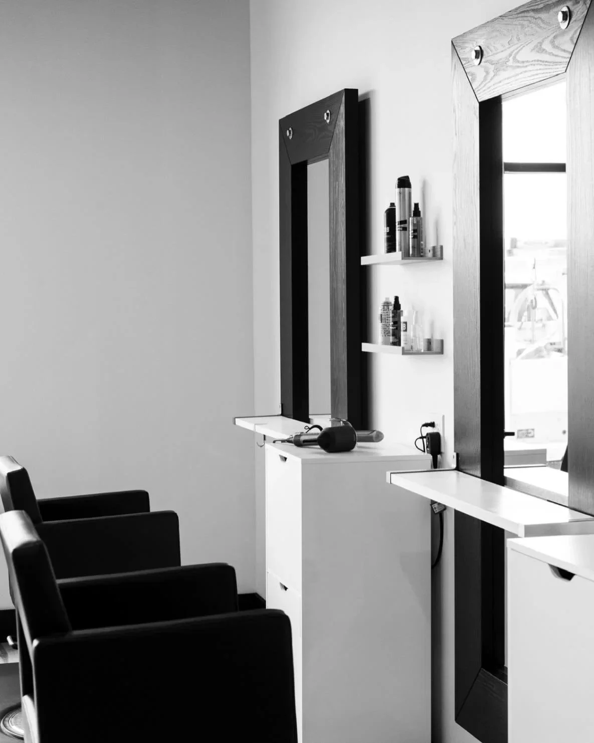 Black and white photo of a salon or barbershop setup with four chairs and two mirrors, with shelves holding products, and a hairdryer on the counter.