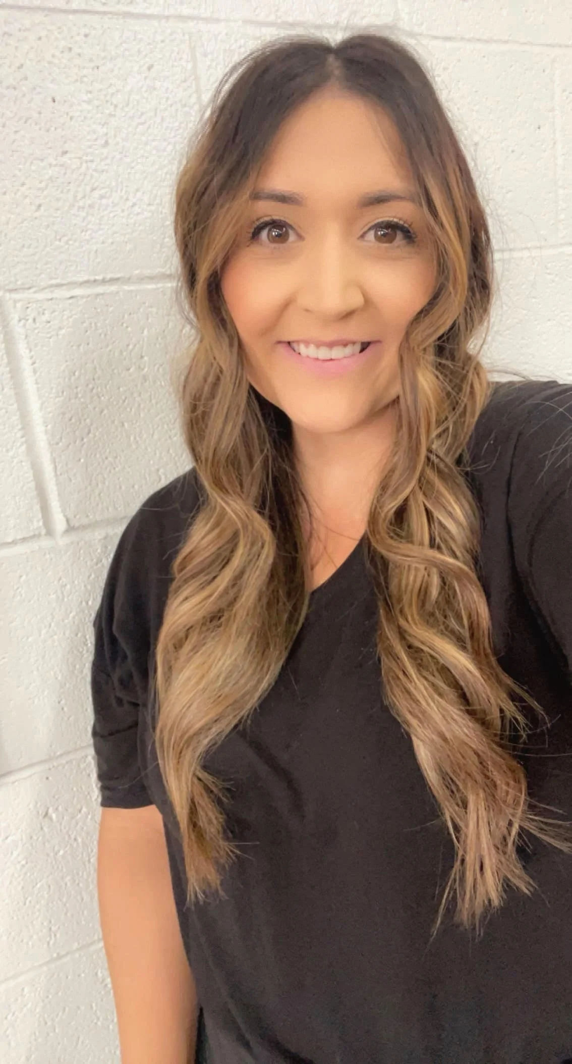A woman with long, wavy brown hair, smiling, wearing a black shirt, standing against a white brick wall.