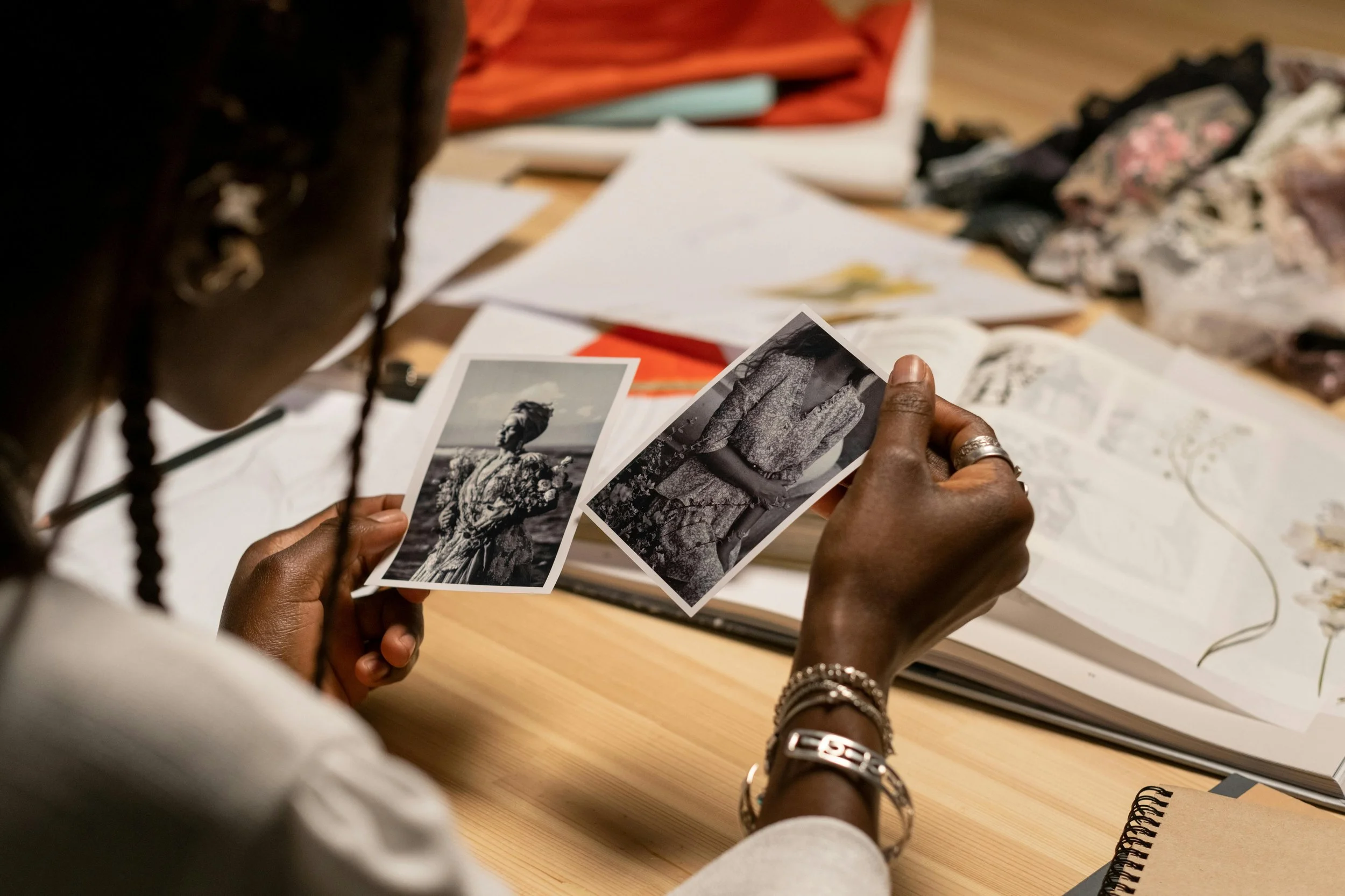Person holding two black and white photographs of a woman, sitting at a wooden desk with papers, sketchbooks, and fabric swatches scattered around.
