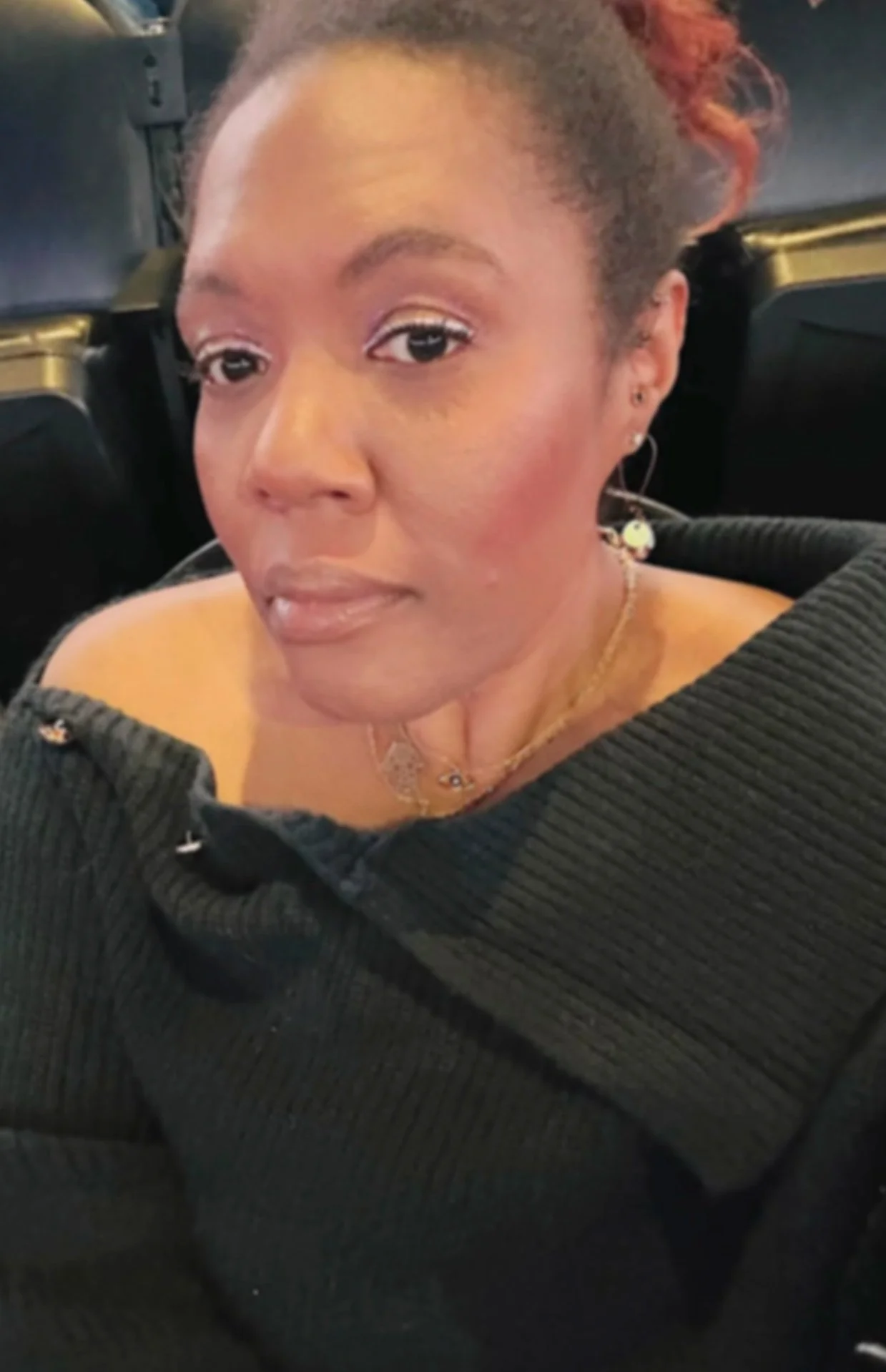 A woman sitting on a plane, wearing a black off-shoulder sweater, with earrings, necklaces, and makeup, looking at the camera.