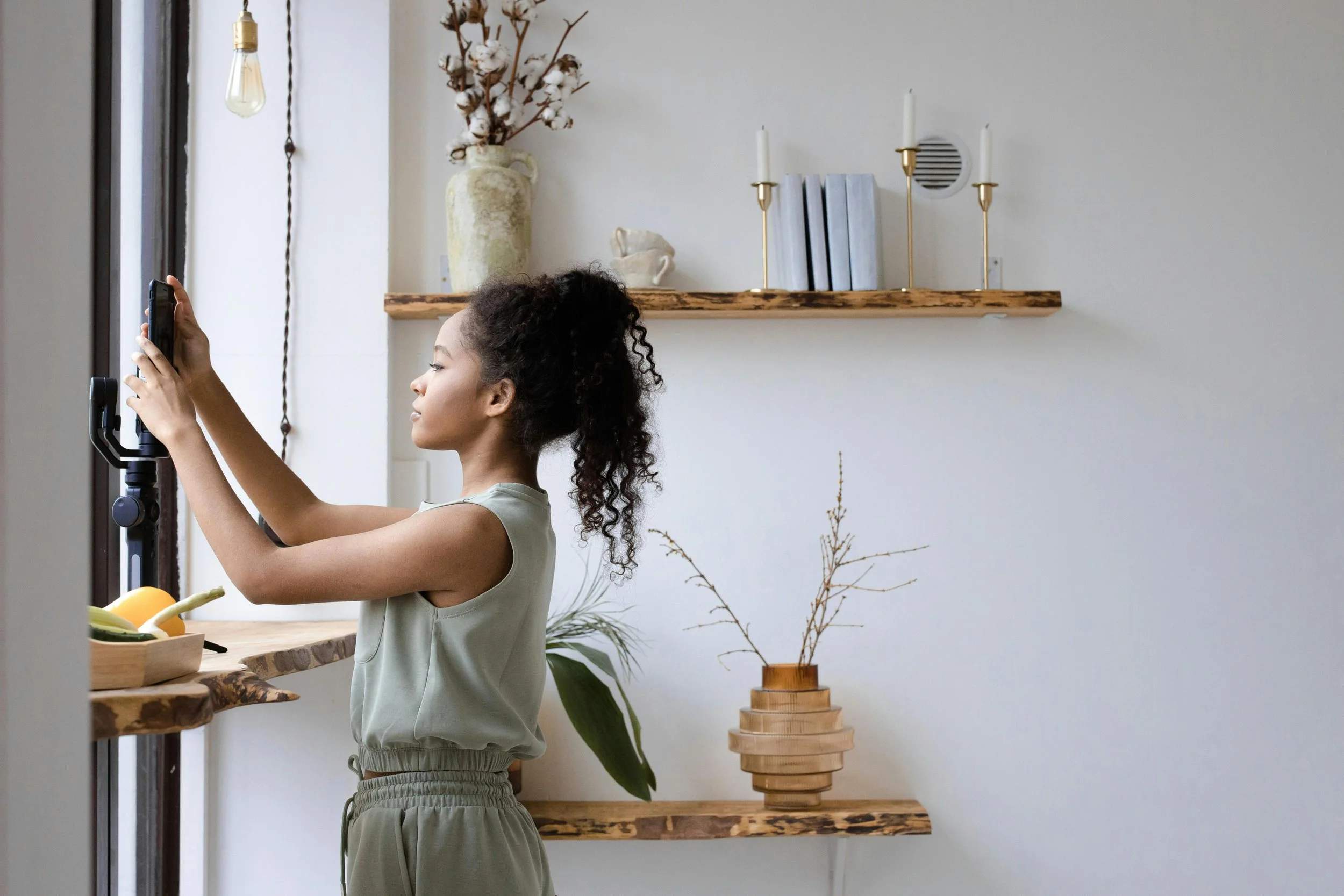 A young girl with curly hair standing near a kitchen window, holding a phone on a selfie stick.