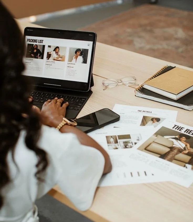 Person working at a desk with a laptop, smartphone, glasses, notebooks, and printed documents.