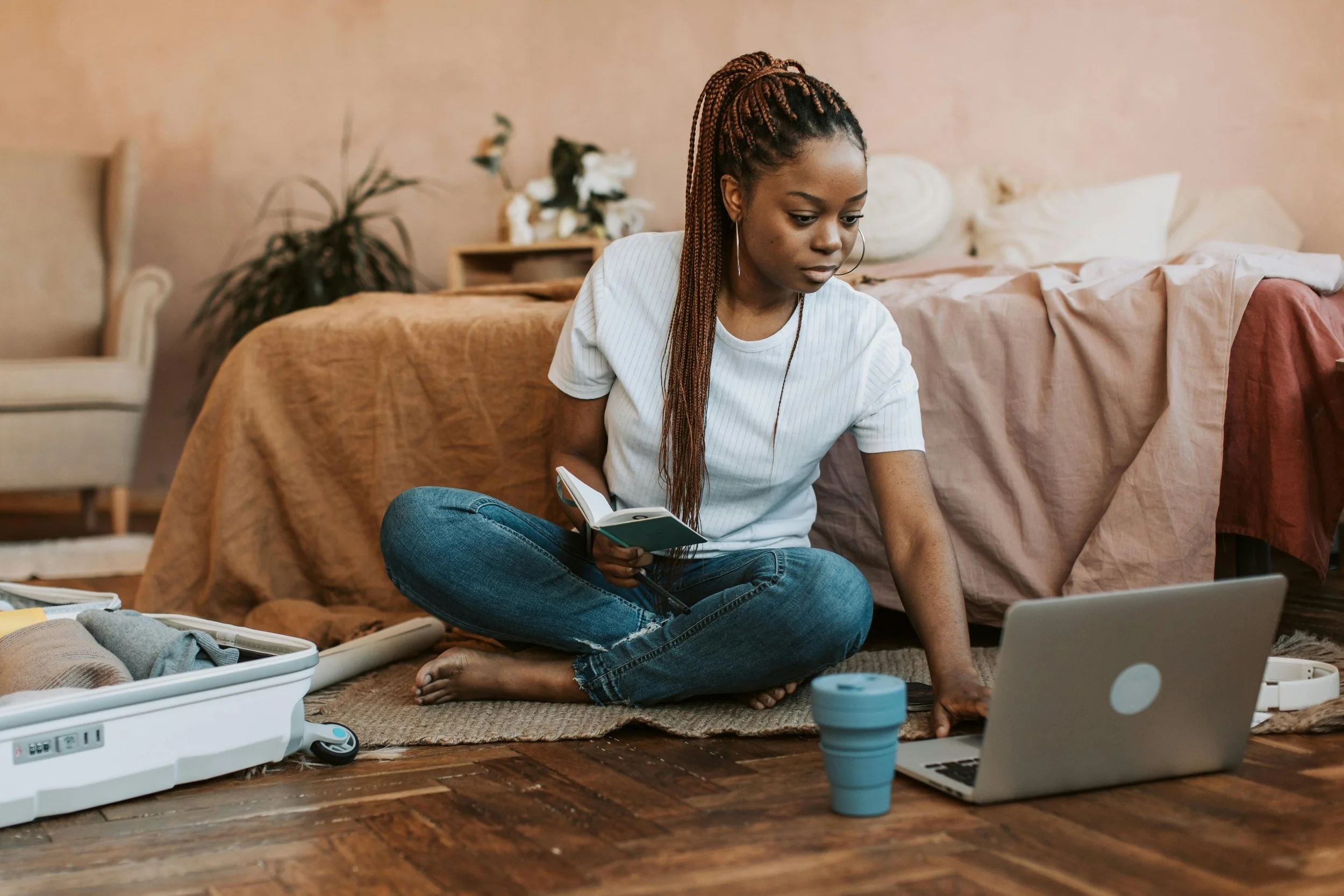 A young woman with braided hair sits on the floor of a bedroom, looking at her laptop with a coffee cup and a small book in her hand. She is dressed casually in a white t-shirt and jeans, surrounded by a suitcase and various items.
