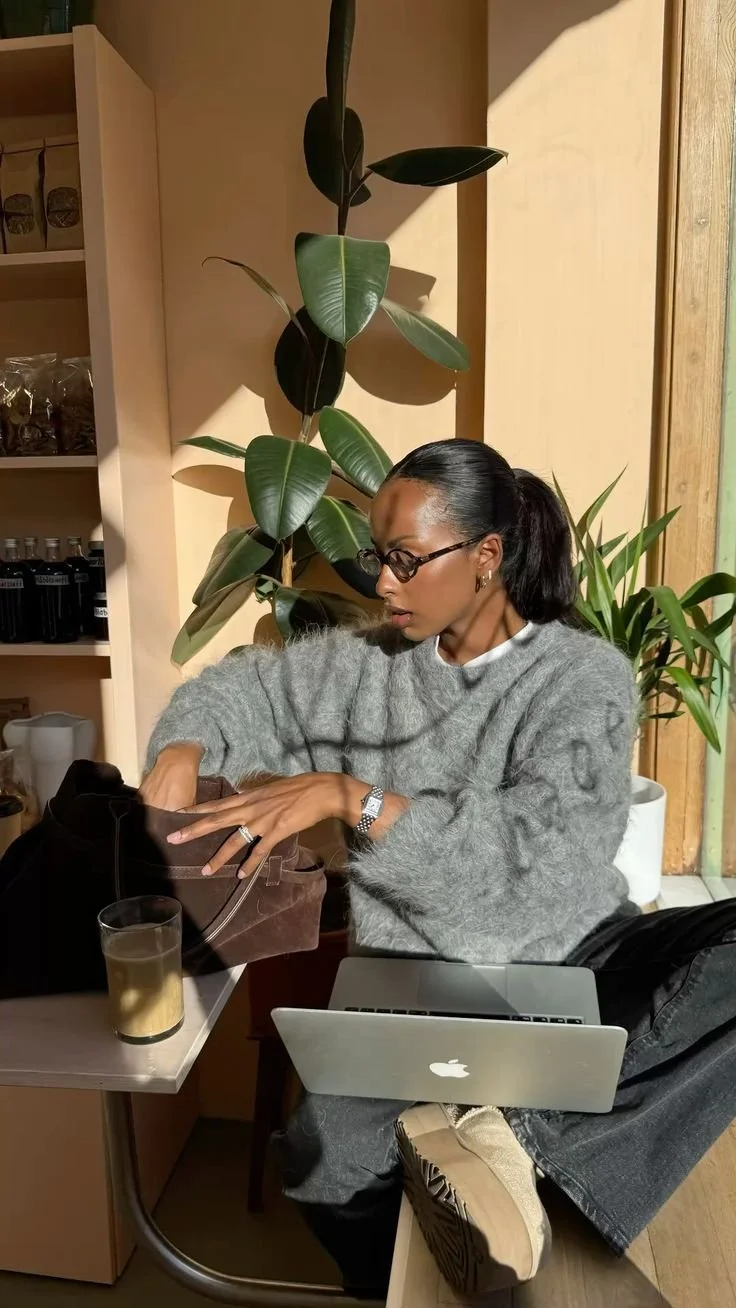A woman with glasses and a ponytail, wearing a gray fuzzy sweater, is sitting at a table with a laptop, reaching into a brown bag. She has a glass of a light-colored beverage, a wristwatch, and a ring. Behind her, there are large green houseplants and wooden shelving with bottles and jars inside a cozy indoor space.