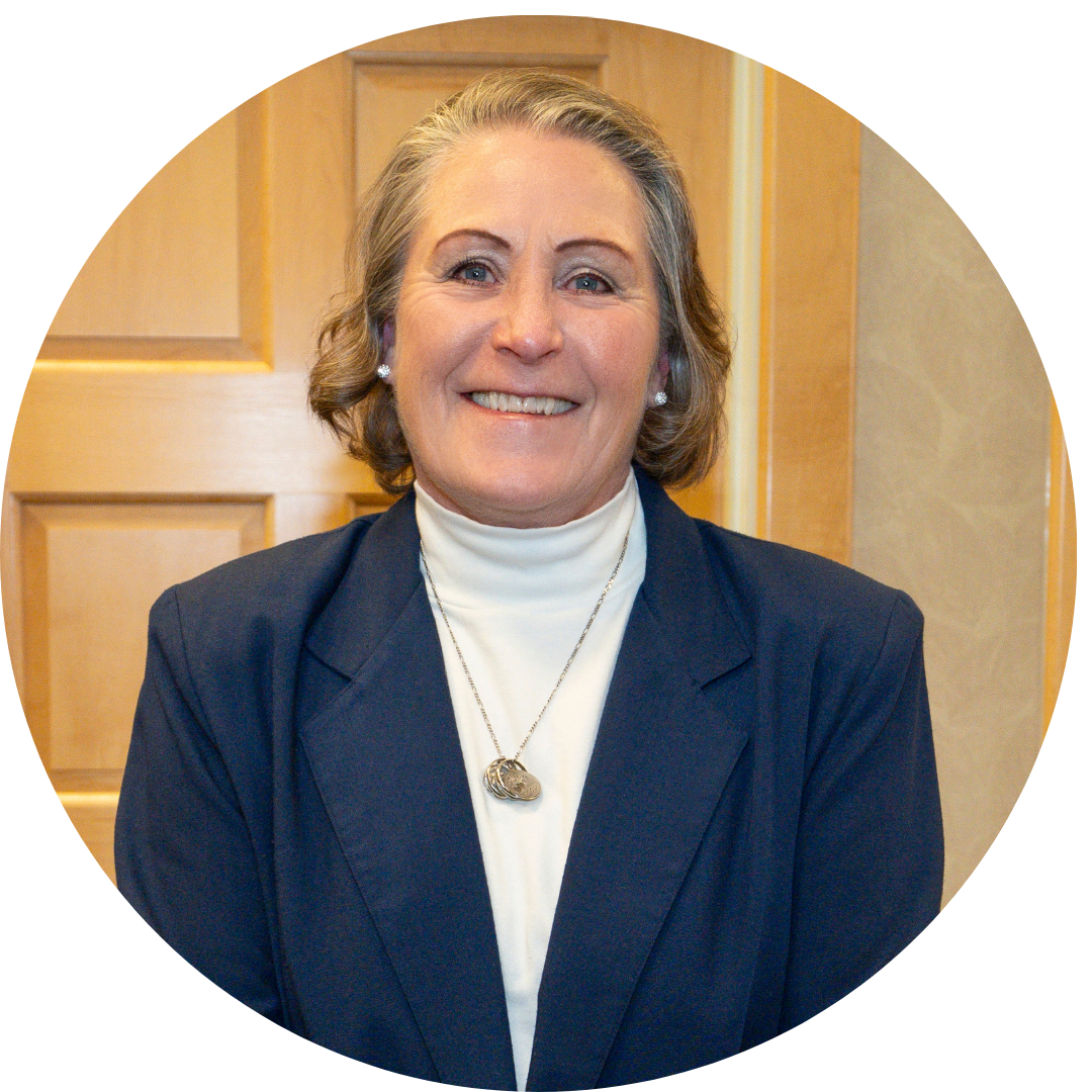 Portrait of a smiling  woman with short gray hair, wearing a dark blazer over a white turtleneck, with a necklace and earrings, standing in front of a wooden door in an indoor setting.