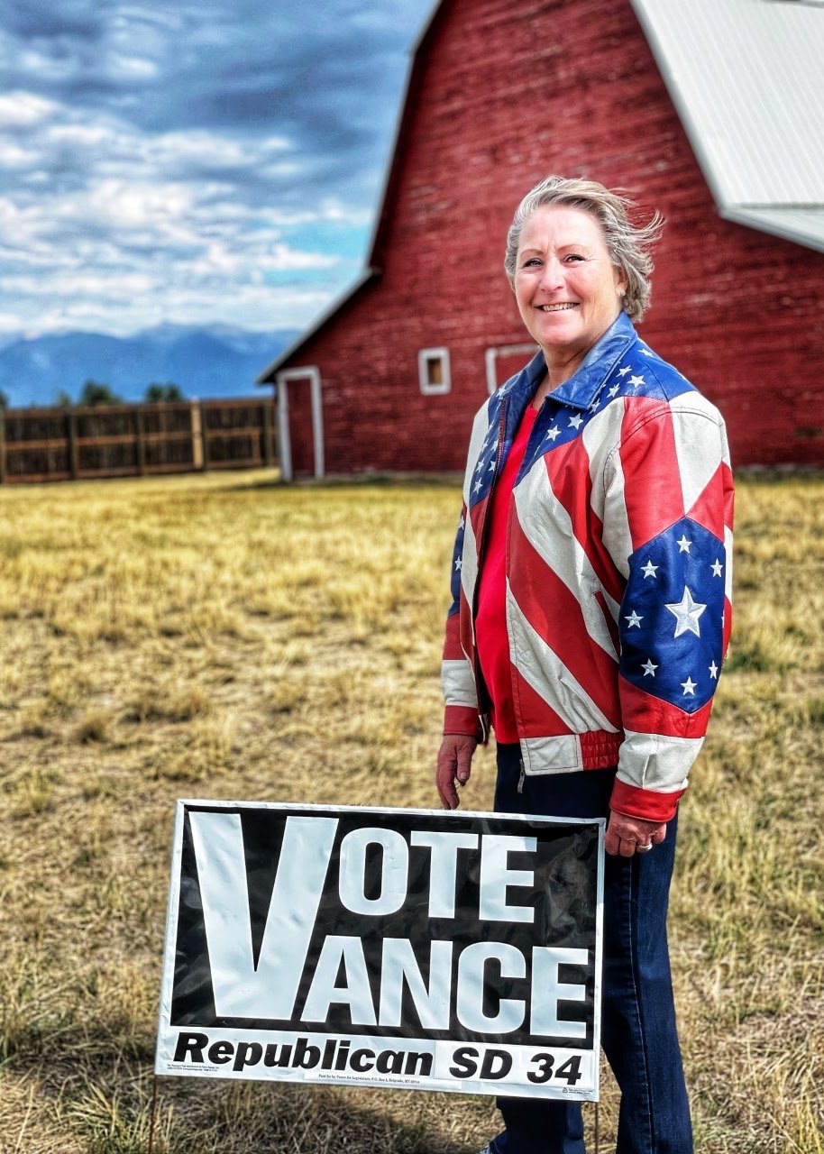 An woman outdoors on a grassy field, smiling, holding a political campaign sign that reads 'VOTE VANCE Republican SD 34'. She wears a jacket with the American flag pattern, and behind her is a red barn and cloudy sky.