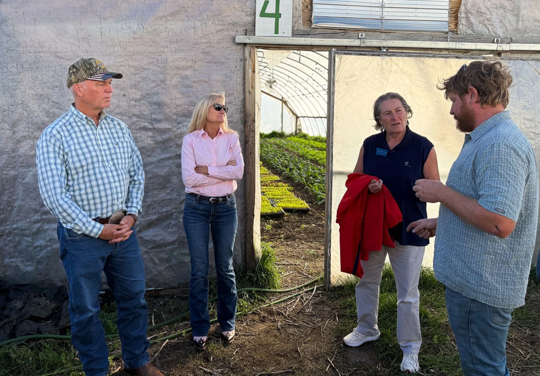 Four people standing inside a greenhouse, with soil and plants visible in the background, engaged in a conversation.