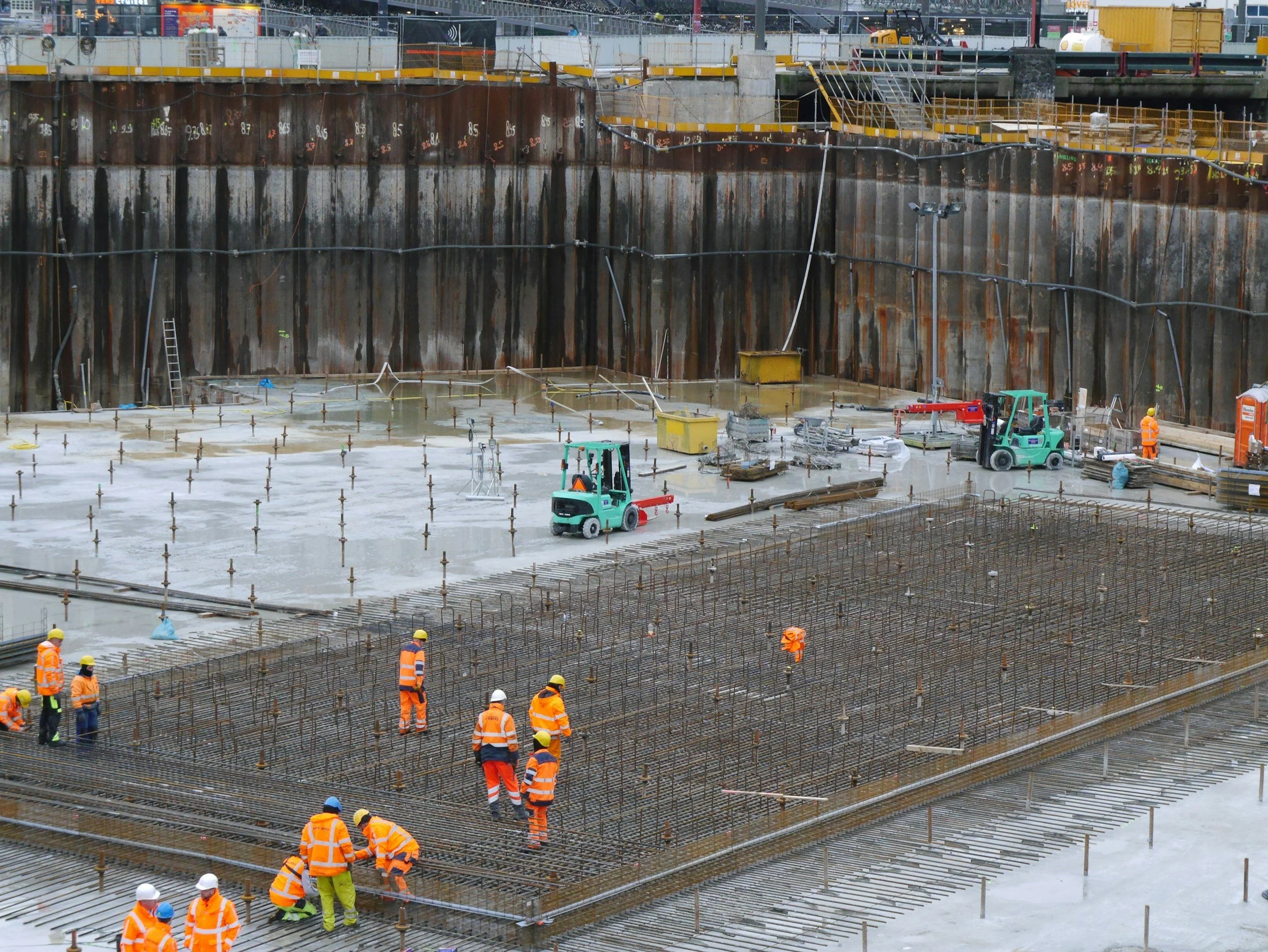 Construction site with workers in orange safety vests and helmets, concrete being poured into rebar framework, and heavy machinery on site.