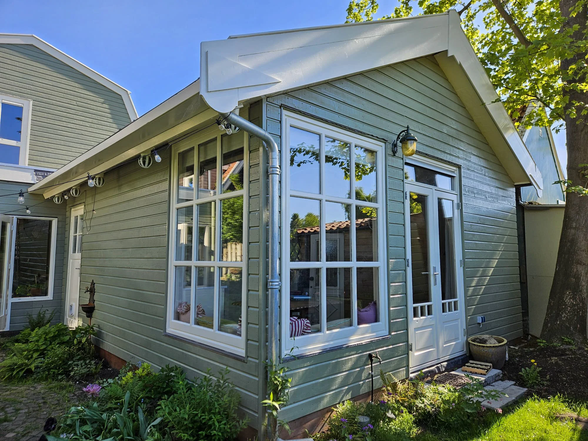 Groene houten huis met grote ramen en een witte deur, rondom een tuin met planten en een grote boom, in de buurt van een blauwe hemel.