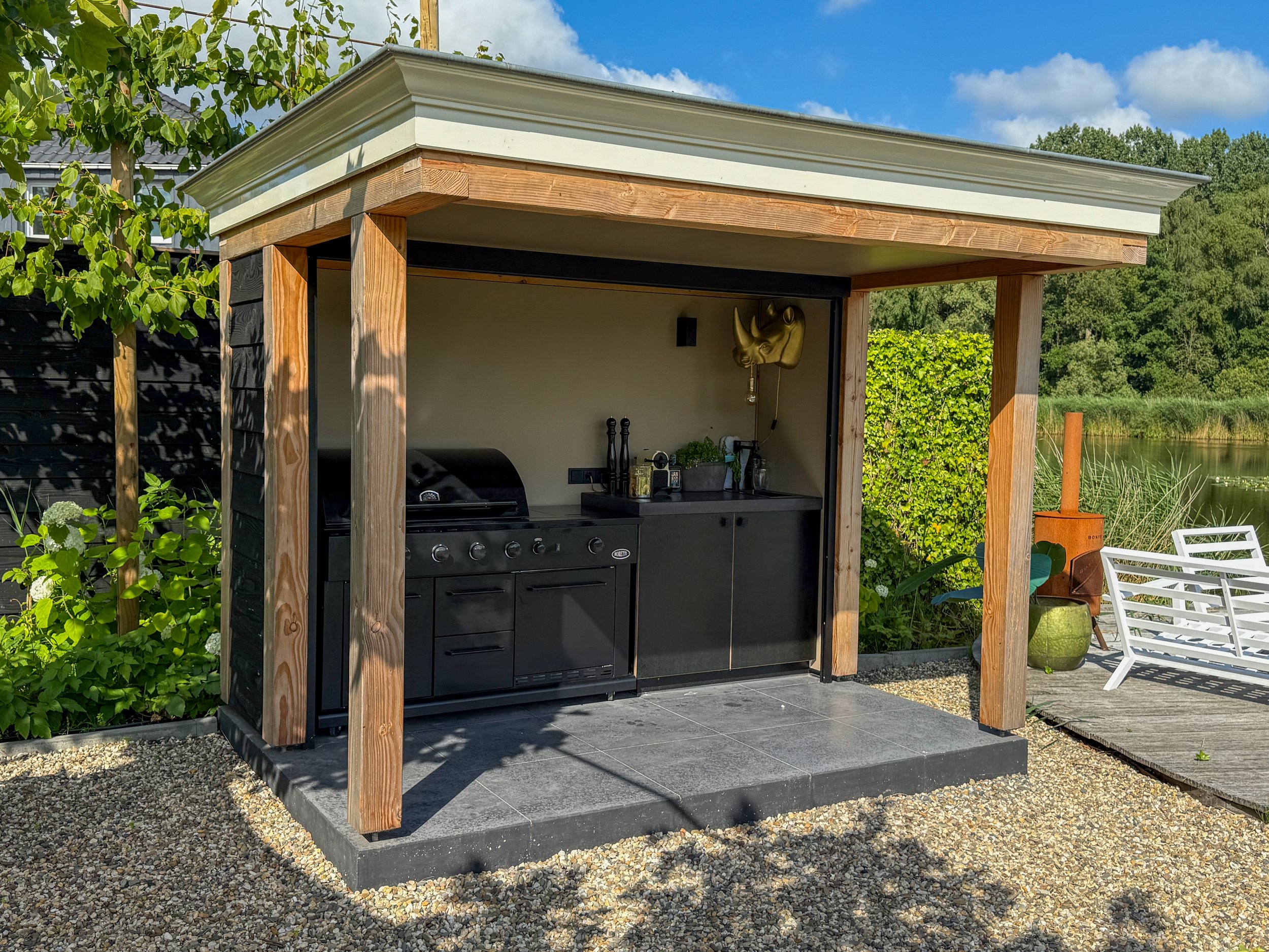 Een buitenkeuken met een barbecue onder een overkapping, omgeven door groen, met een terras en een meer op de achtergrond.
