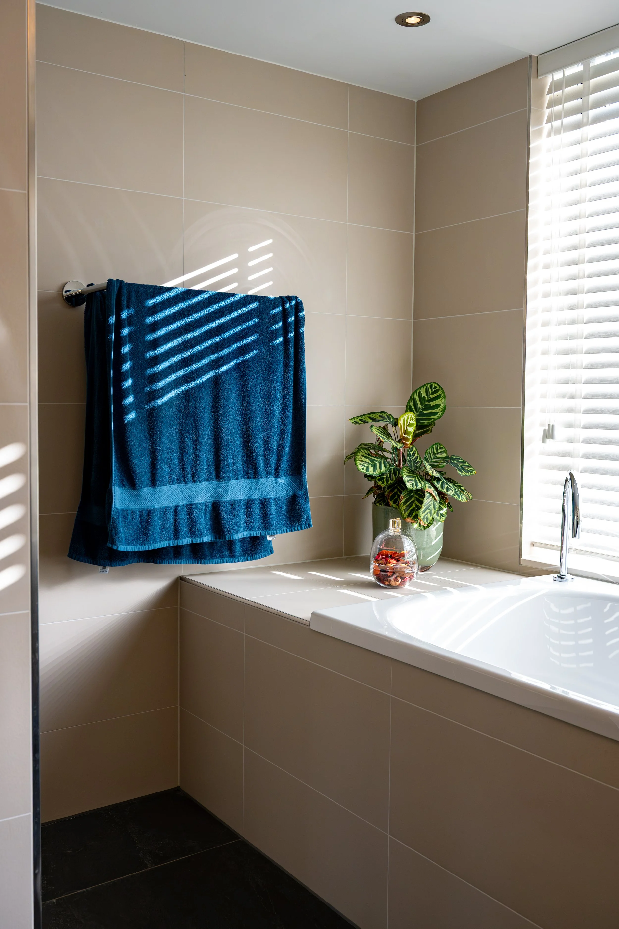 Een moderne badkamer met beige tegels, een wit bad, een raam met witte jaloezieën, een groene plant en een glazen pot met decoratieve voorwerpen.