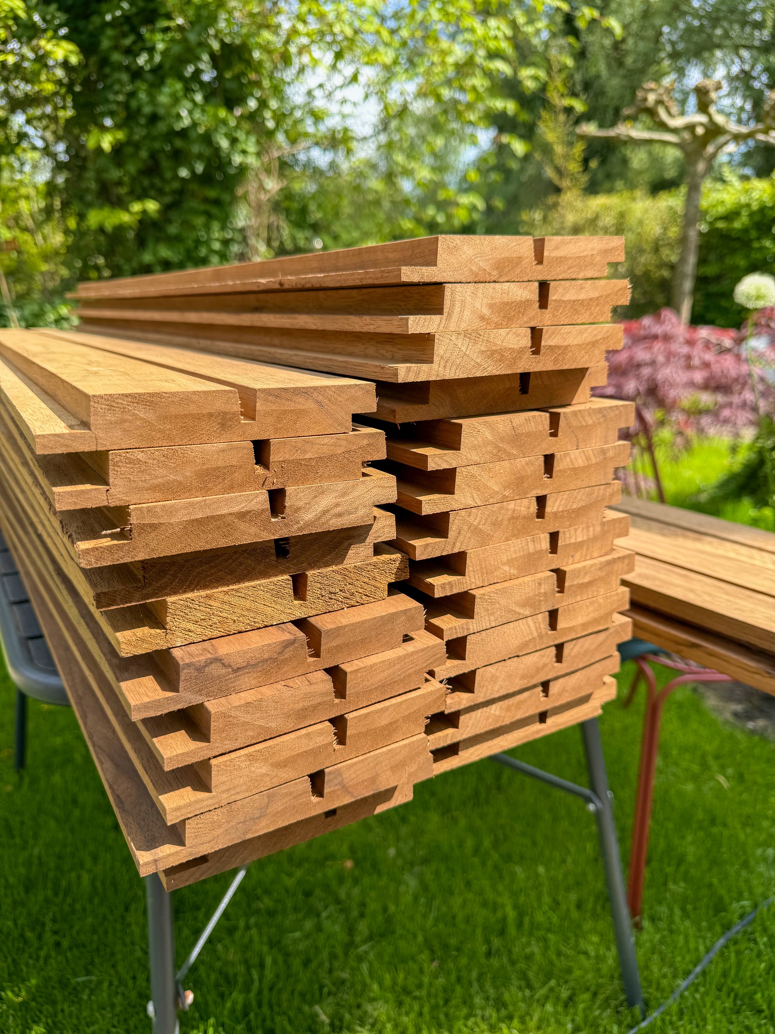 Een stapel houten latten ligt op een tafel in een tuin, met groene bomen en struiken op de achtergrond.