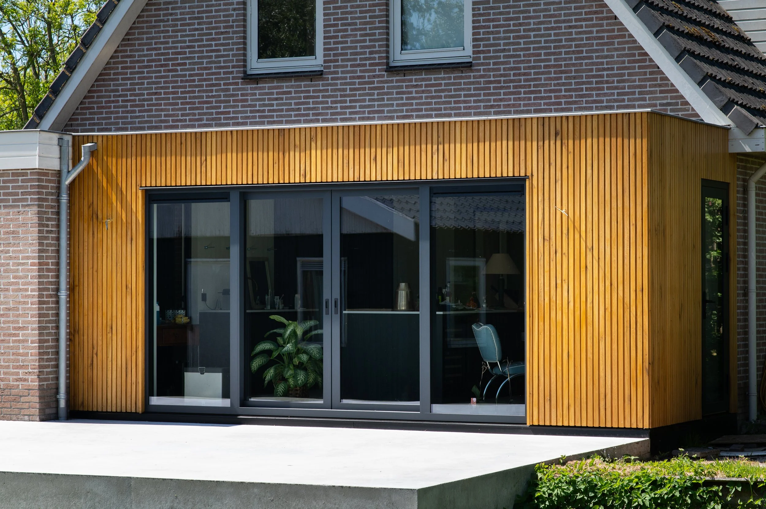 Modern huis met een houten uitbouw en grote glazen deuren, een plant binnen zichtbaar.