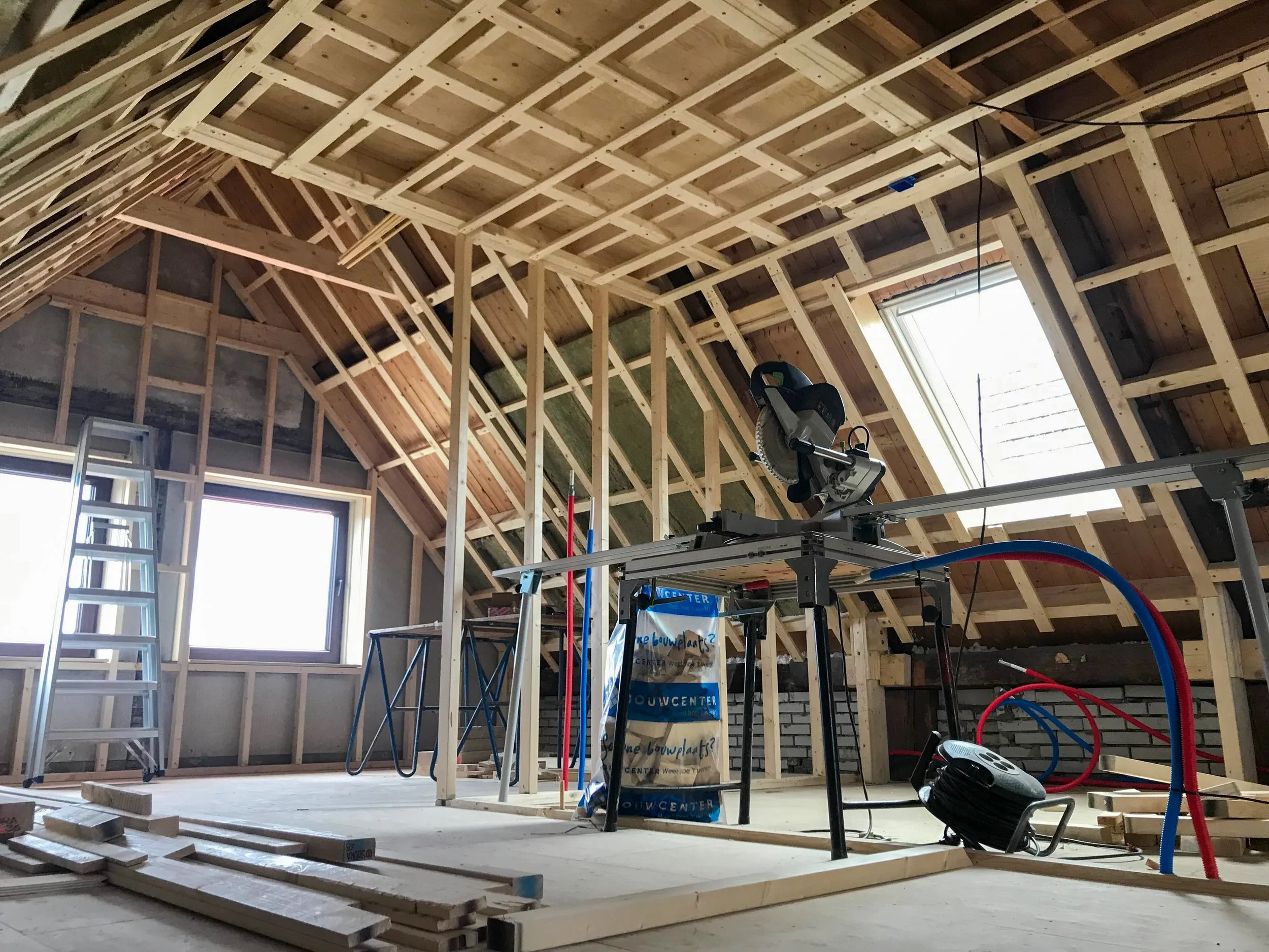 Binnenkant van een zolder in aanbouw met houten raamwerk en een grote schuifdakraam. Er staat een zaagmachine op een werkbank, en er zijn ladders en bouwmaterialen rondom.