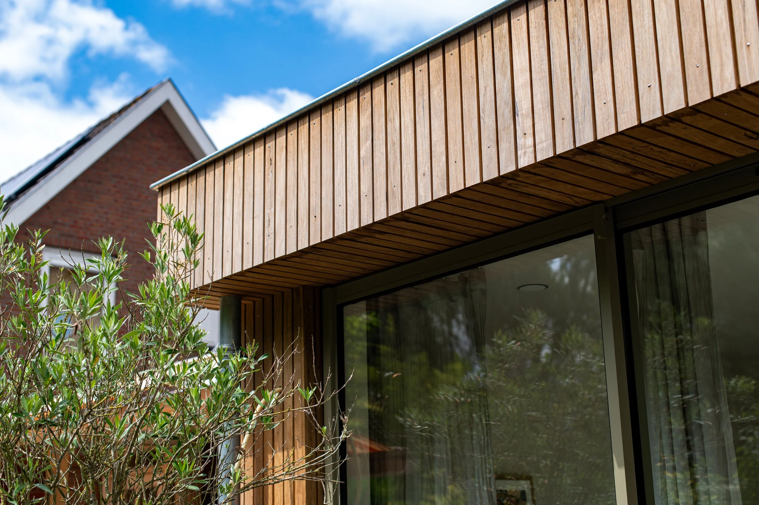 Houten gevel van een modern huis met grote glazen ramen en groene planten voor het raam, tegen een blauwe lucht met wat wolken.