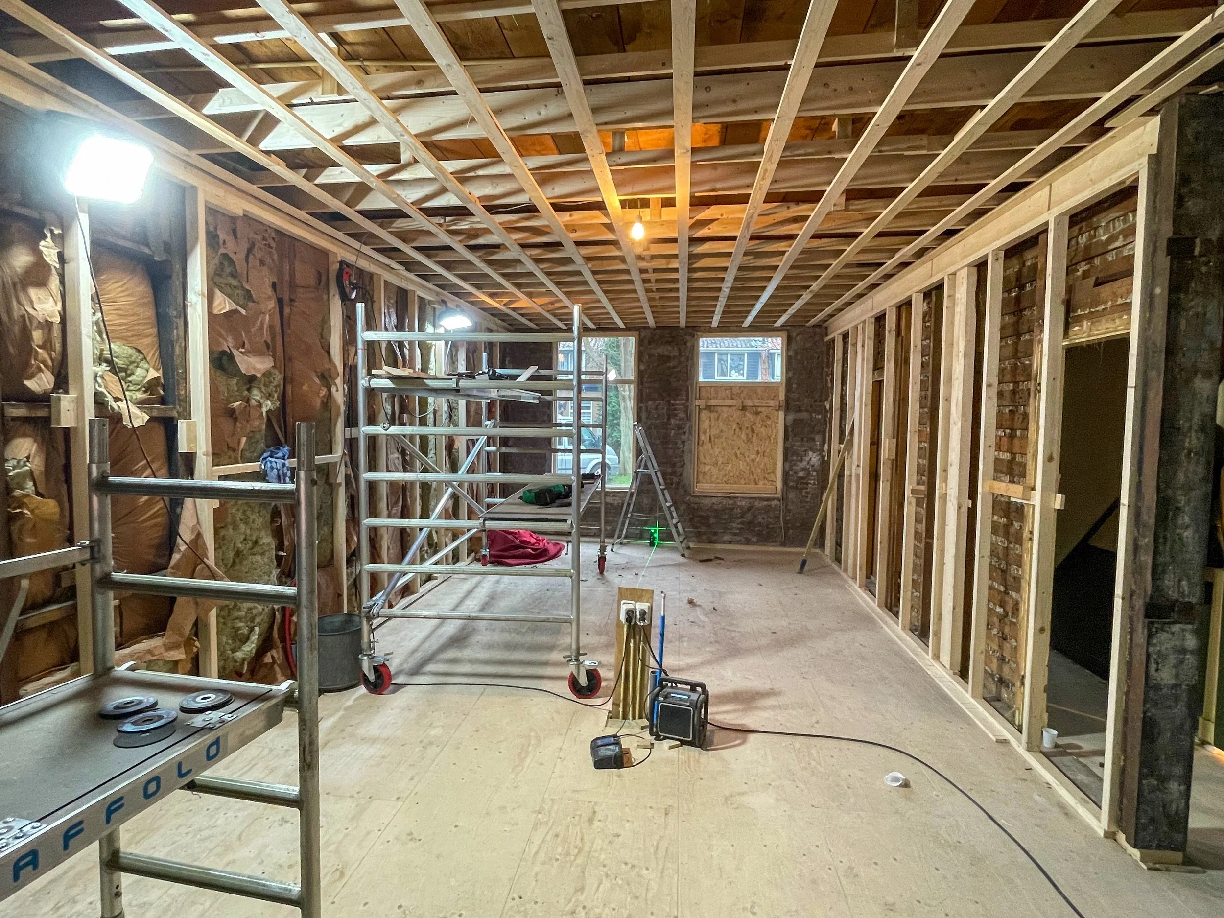 Binnenkant van een kamer in aanbouw met houten wanden en plafond, scaffolding, gereedschap en bouwmaterialen, ramen en een deur afgewerkt.