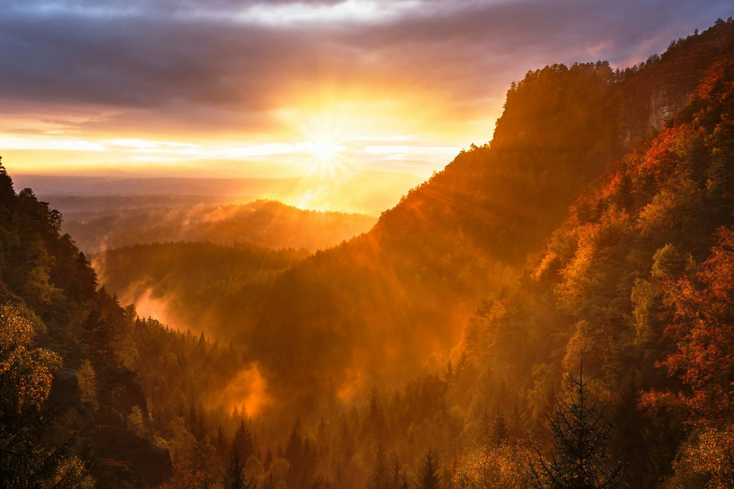 Sunrise over a mountain valley with mist and dense forest, displaying autumn colors.