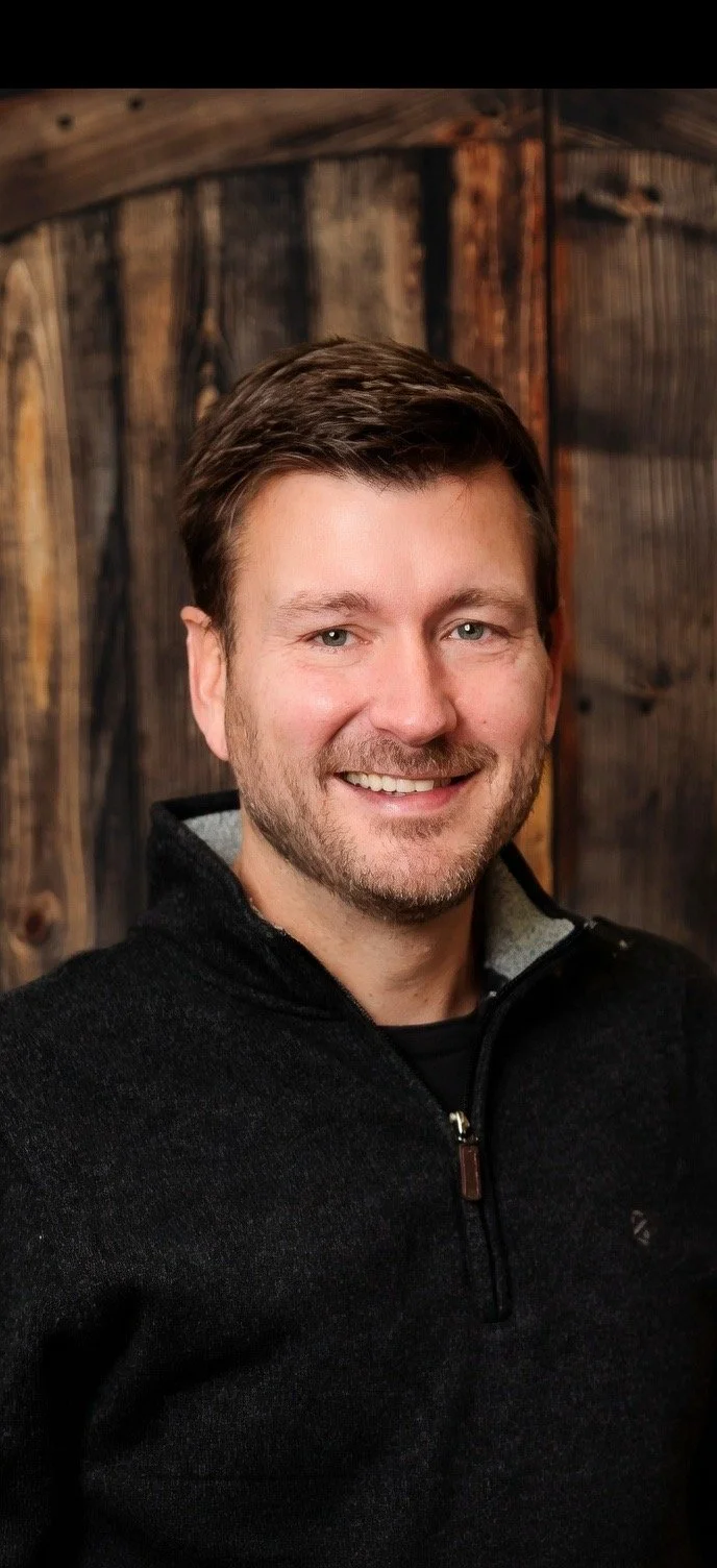 Portrait of a smiling man with short brown hair, a beard, wearing a black zip-up sweater, against a wooden background.