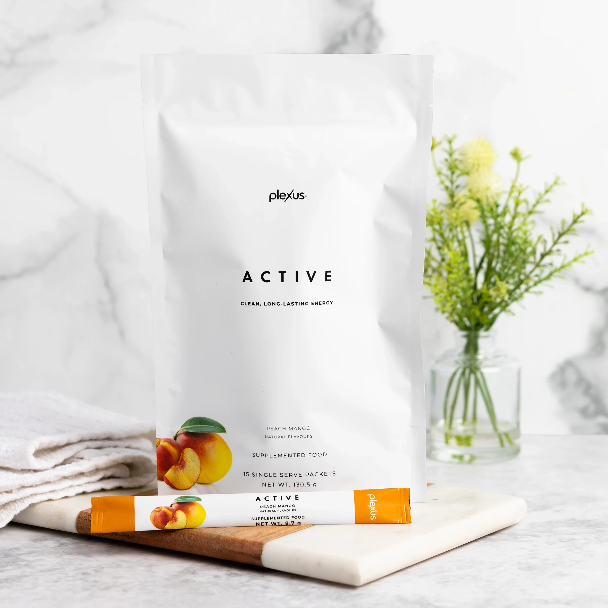 White packaging of Plexus Active Peach Mango supplement on a marble surface with a white towel, wooden board, and a glass jar with yellow flowers in the background.