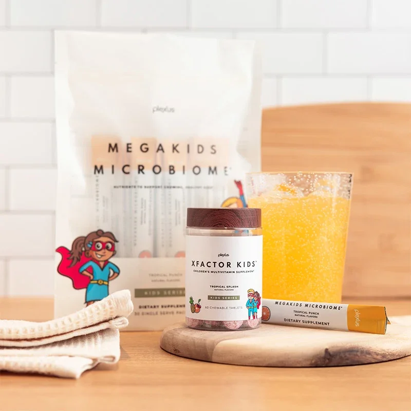 Children's multivitamin supplement bottle, chewable tablets, a stick of dietary supplement, a glass of orange juice, and a bag of MegaKids Microbiome probiotic powder on a wooden surface against a white tile wall.