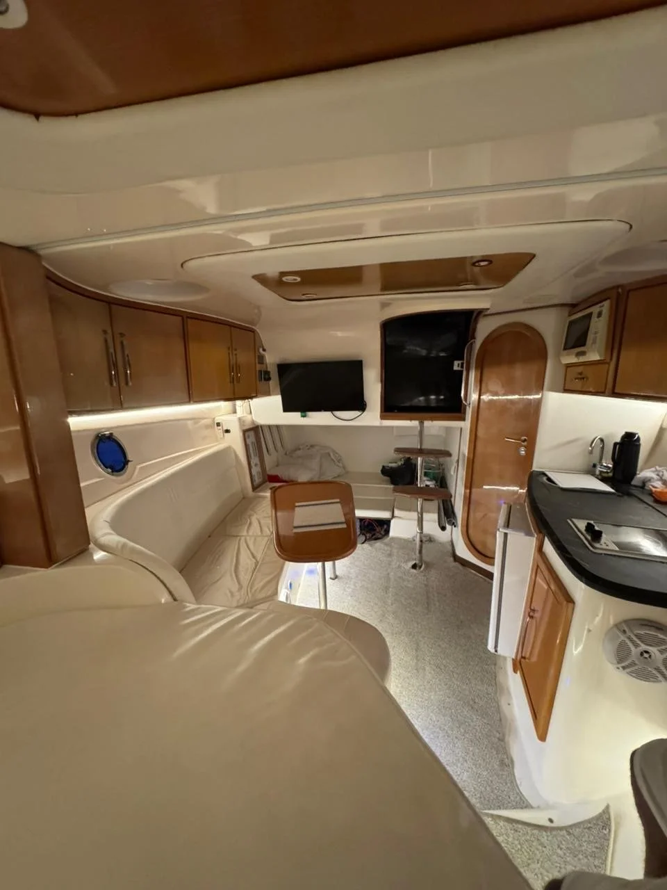 Interior of a boat or yacht cabin showing a cozy living area with a cream-colored sofa, a curved wooden table, a flat-screen TV, wooden cabinets, and a compact kitchenette with a sink, coffee maker, microwave, and other appliances.