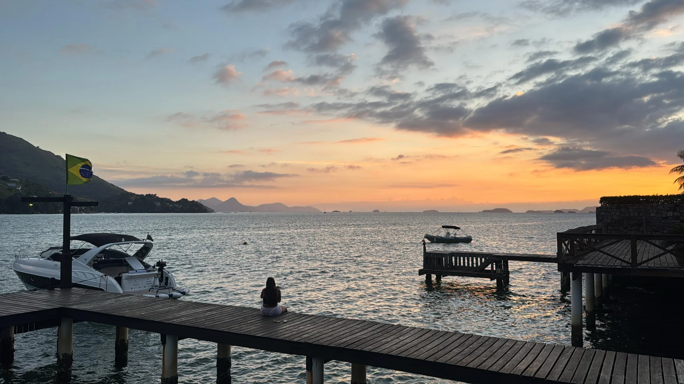 A person sitting on a wooden pier by the water during sunset, with boats and a Brazilian flag on a pole, hills, and islands in the background.