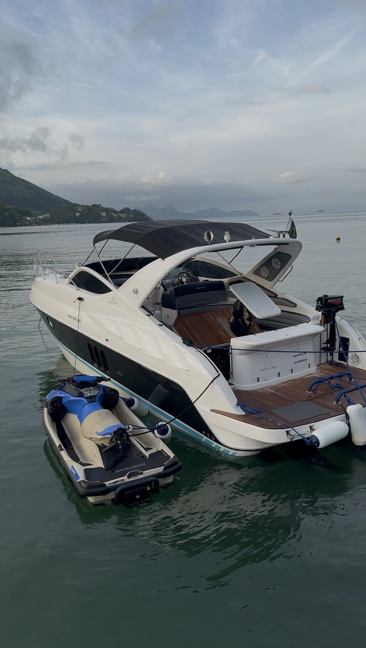 A white yacht with a black canopy docked in calm water near a small inflatable boat. The background features mountains, a cloudy sky, and distant islands.