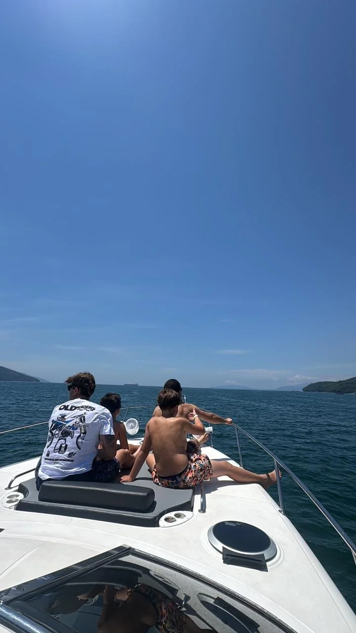 A group of children sitting on the front of a boat, enjoying a sunny day on the water, with a view of a distant shoreline and a clear blue sky.