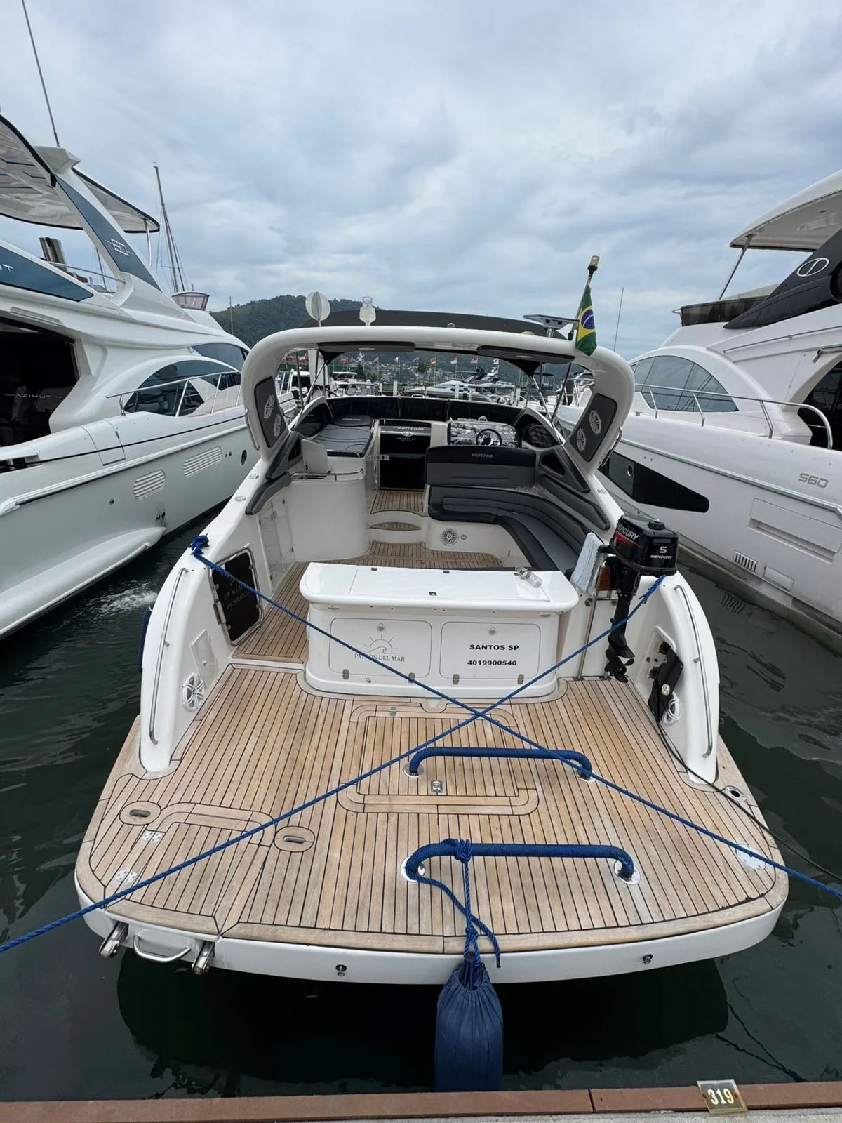 View of a white motor yacht at a marina with other large boats on each side, cloudy sky, and a flag in the background.