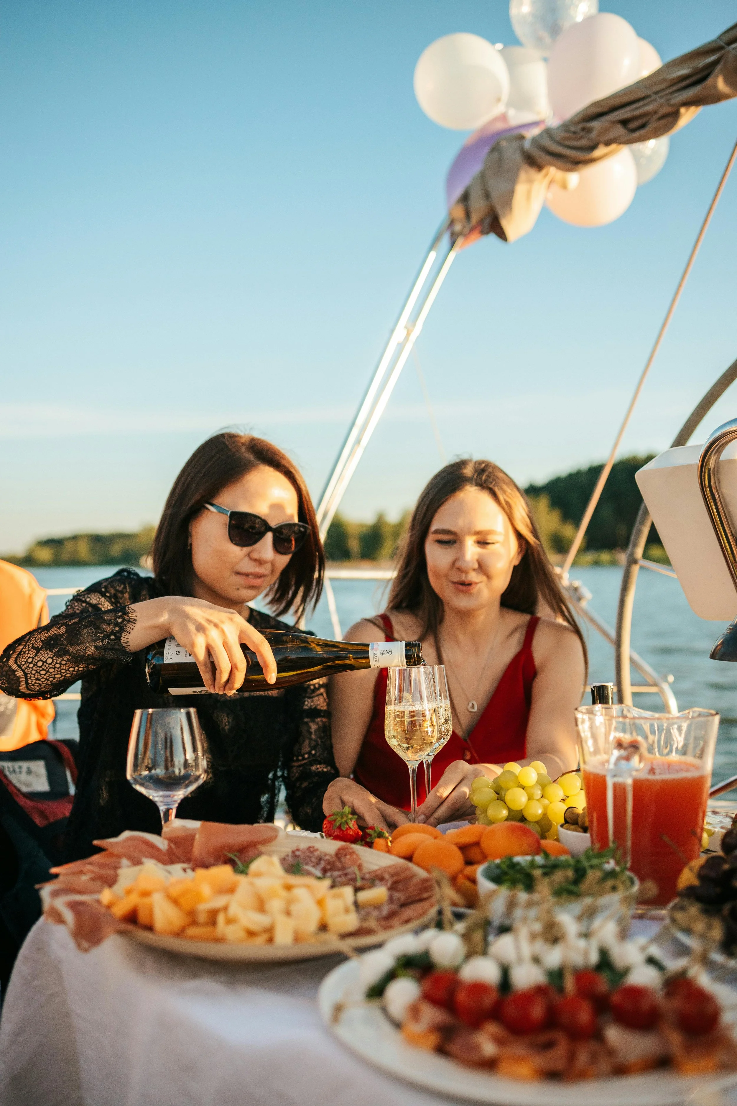 Two women enjoying an outdoor gathering on a boat, pouring wine and surrounded by a spread of food including cheese, grapes, meats, and vegetables, with water and trees in the background.