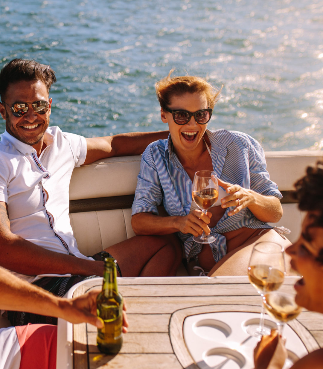 Group of friends smiling and laughing on a boat near the water, enjoying drinks and sunshine.