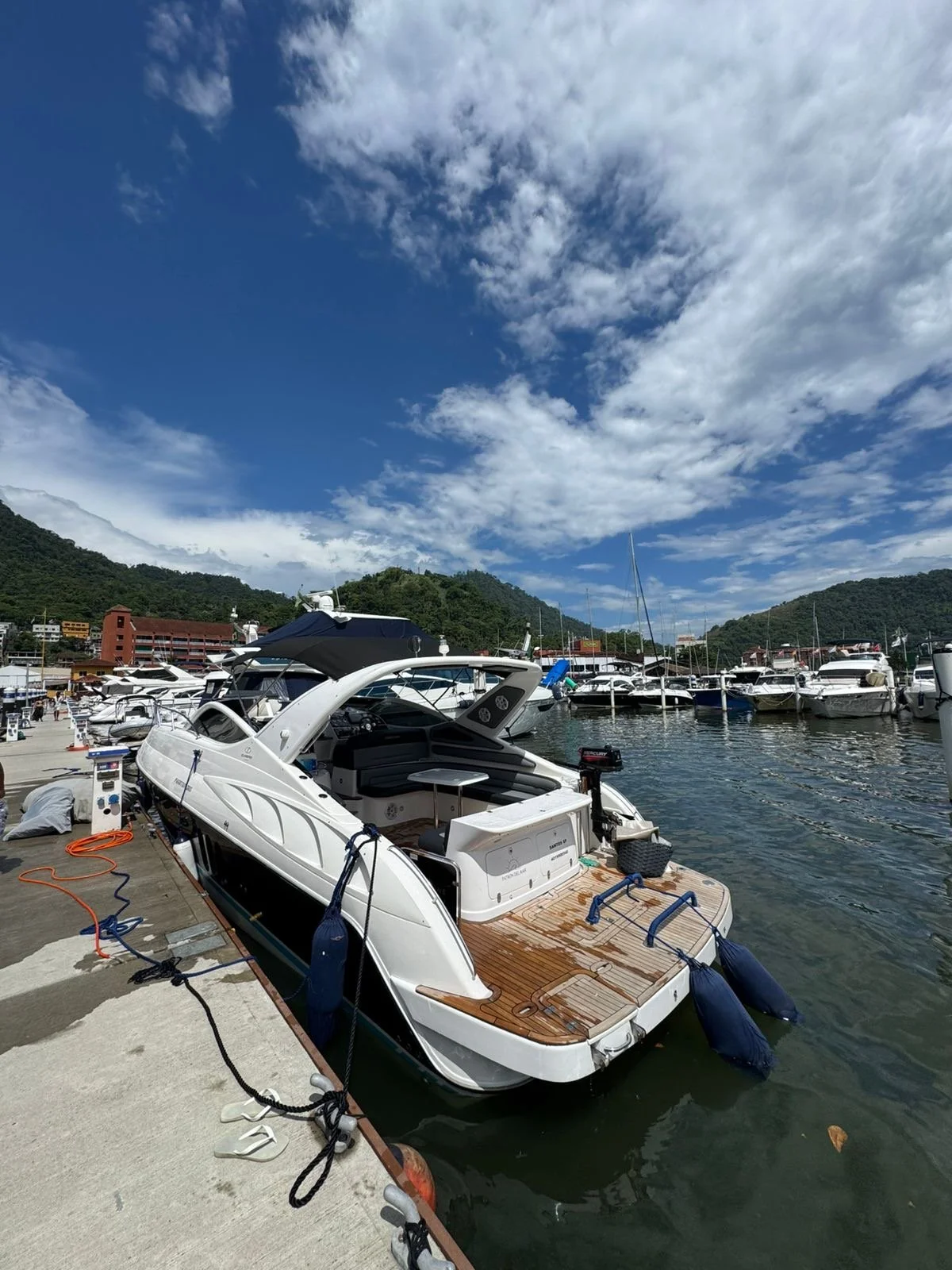 A marina with a white yacht docked at the pier, surrounded by other boats and yachts, with green hills in the background and partly cloudy blue sky overhead.