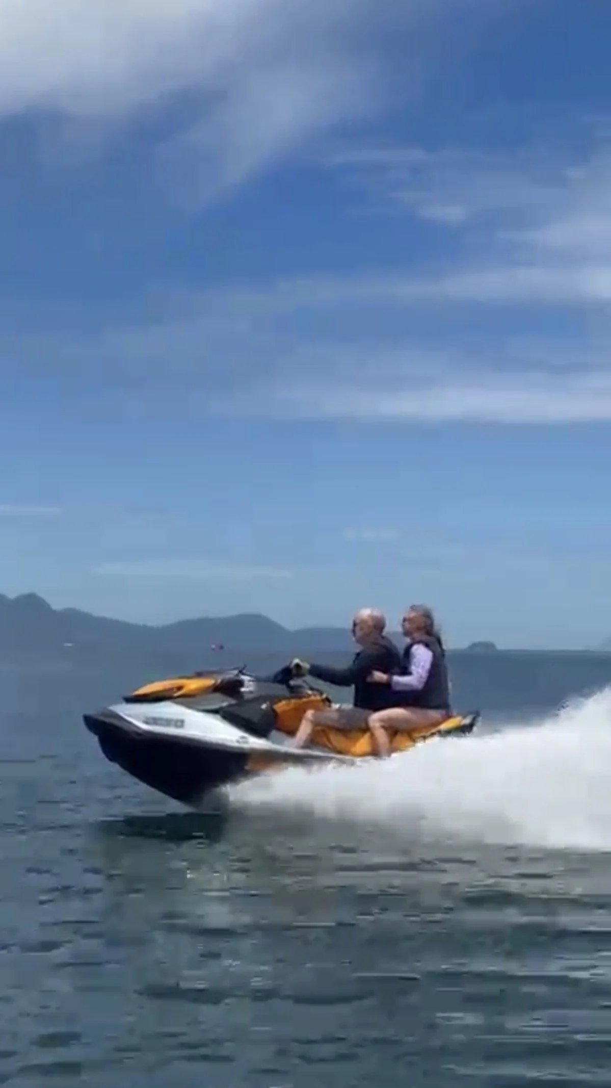 Two people riding a yellow jet ski on the water with mountains in the background and a blue sky with some clouds.