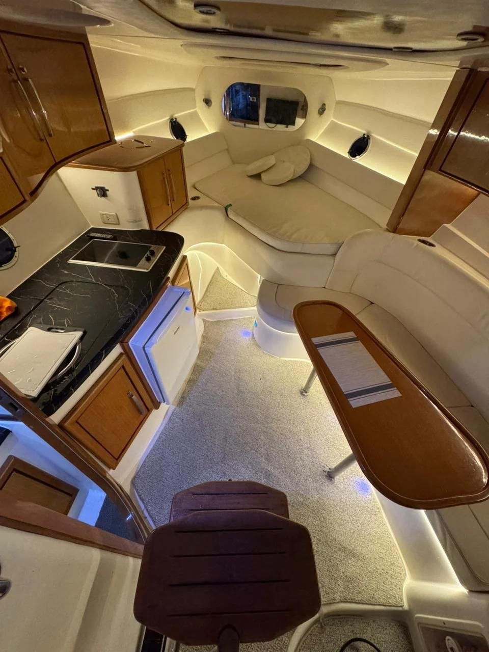 Interior of a yacht showing a kitchenette, a bed, and a seating area with a round table, wooden cabinets, a mini refrigerator, and LED lighting along the ceiling and floors.