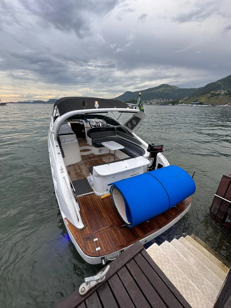 A white yacht docked at a wooden pier on a lake with mountains and cloudy sky in the background.
