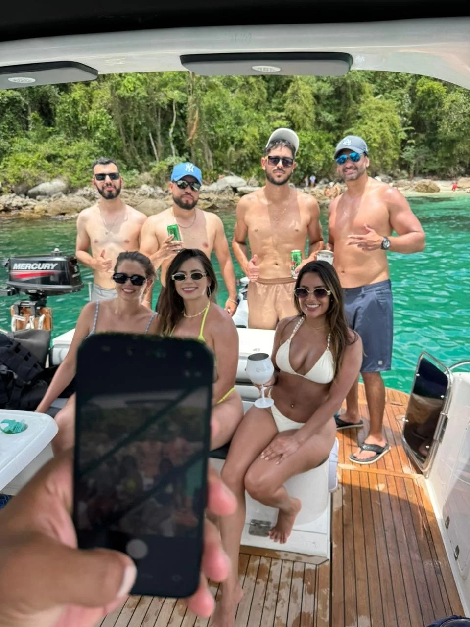 A group of seven friends, including three women in swimsuits and four men in swim trunks, pose on a boat near a green, forested shoreline with rocks. They are smiling and holding drinks, with some wearing sunglasses and hats, enjoying a sunny day on the water.