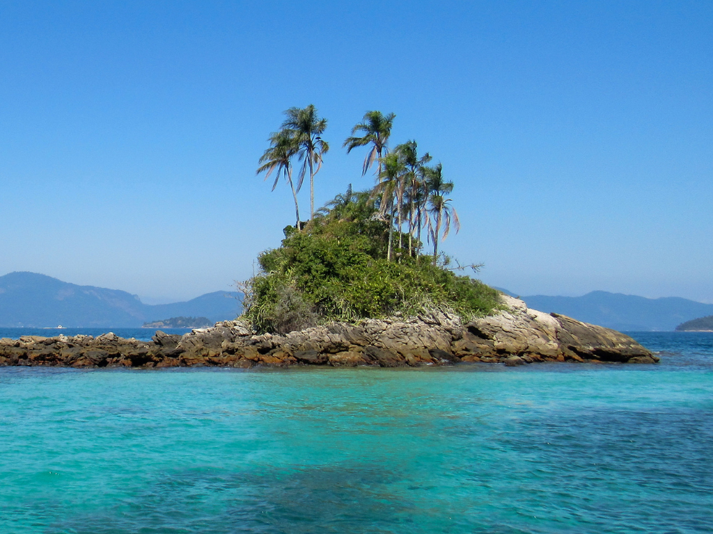 Small rocky island with palm trees surrounded by turquoise water, with blue sky and distant mountains in the background.