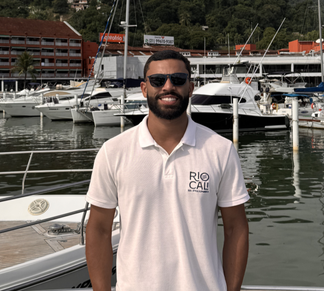 A man with a beard and sunglasses smiling in front of a marina with boats and yachts, hills, and buildings in the background.