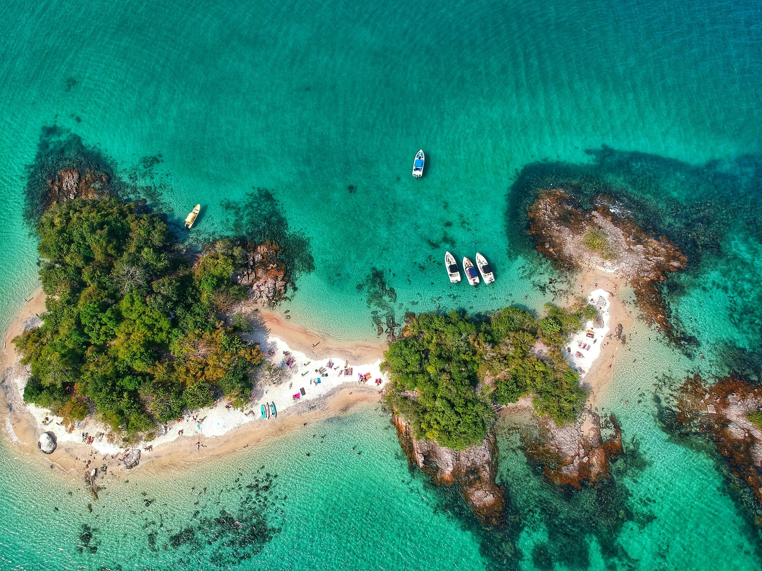 An aerial view of a tropical beach with turquoise water, green vegetation, and several boats anchored near rocky formations and the sandy shoreline.