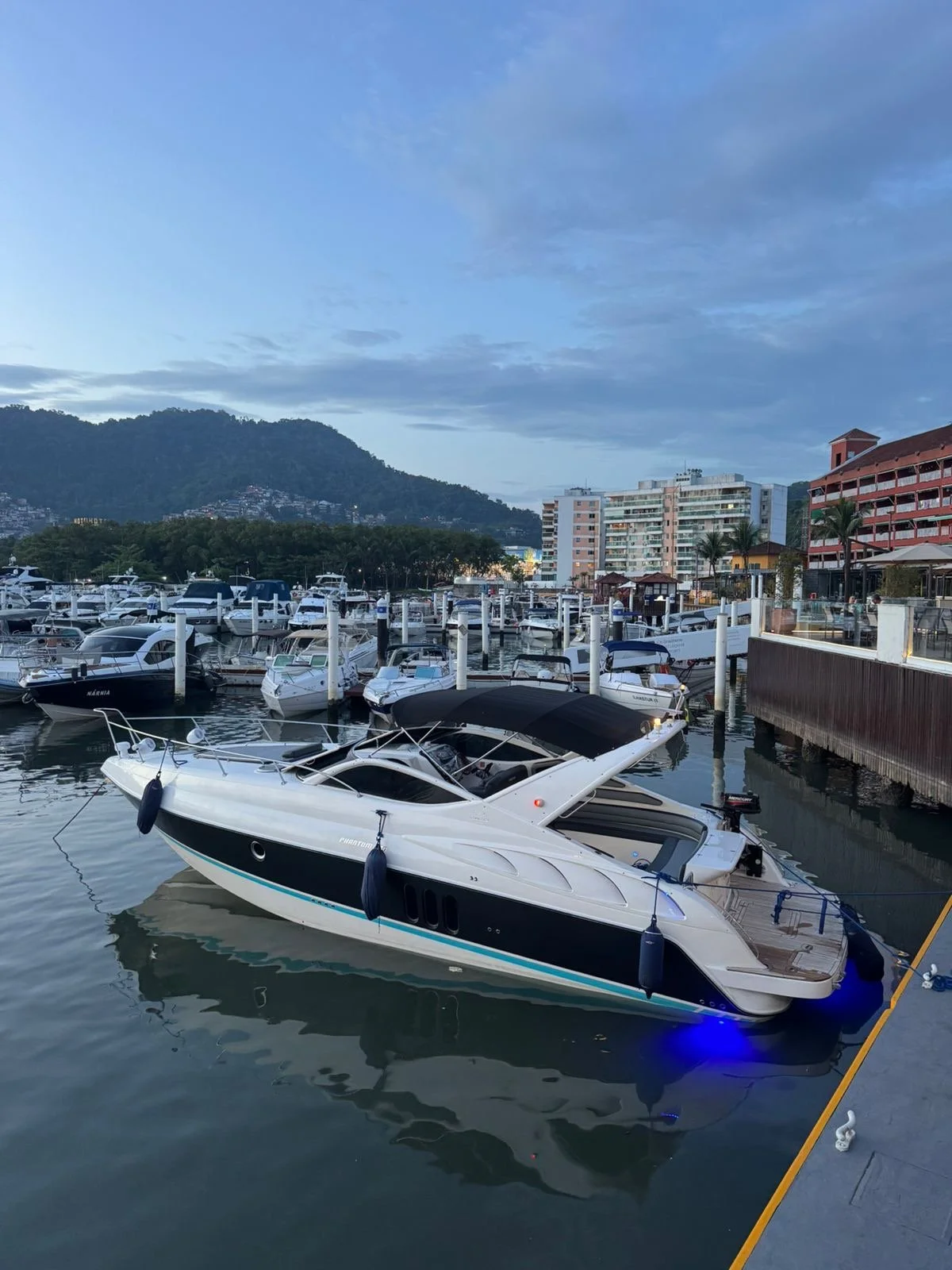 A marina with multiple yachts docked, a large white yacht in the foreground, water, buildings, trees, hills, and cloudy sky in the background.