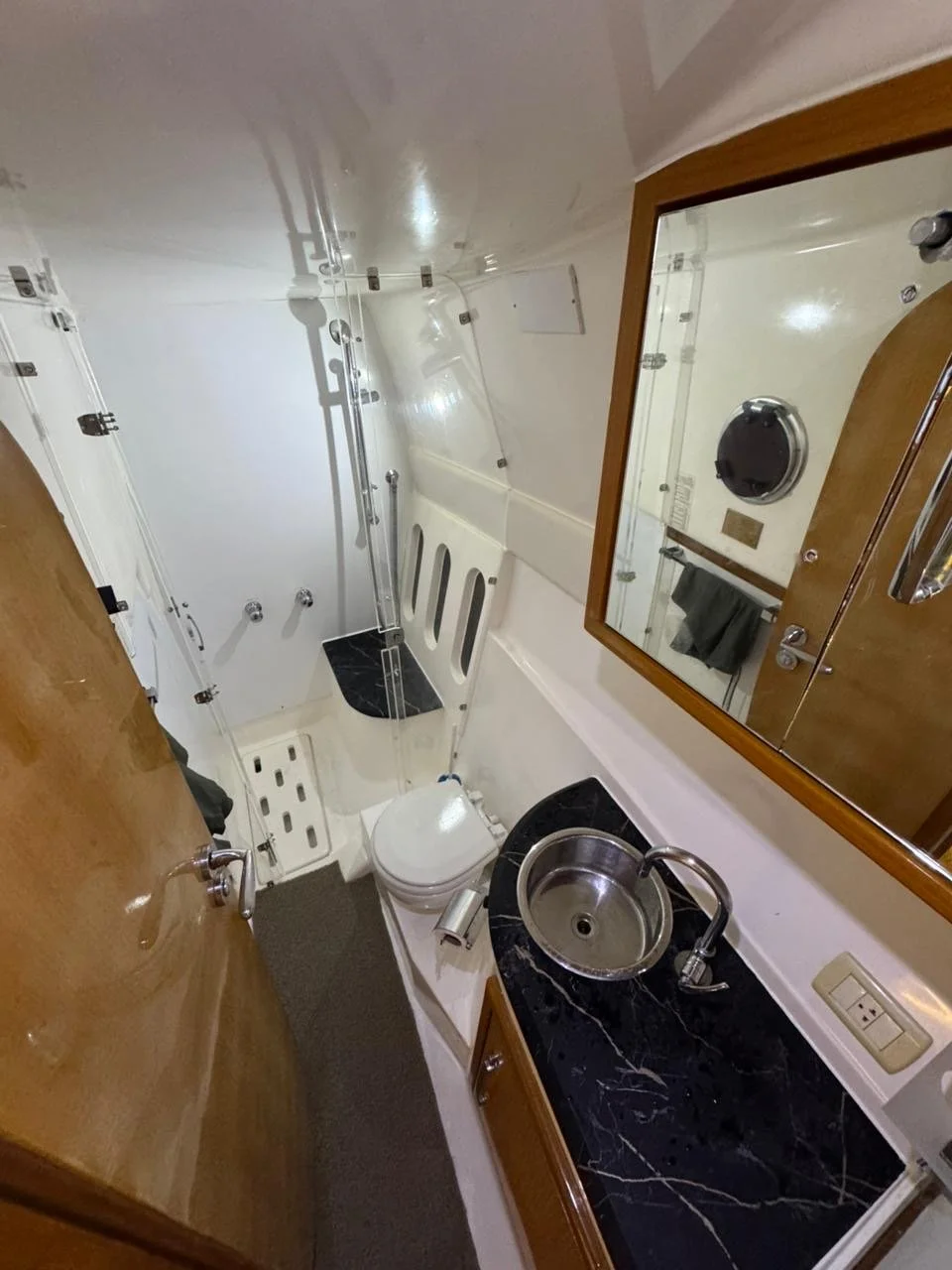 Yacht bathroom with a small round stainless steel sink, black marble countertop, mirror, wooden cabinet, toilet, and enclosed shower.
